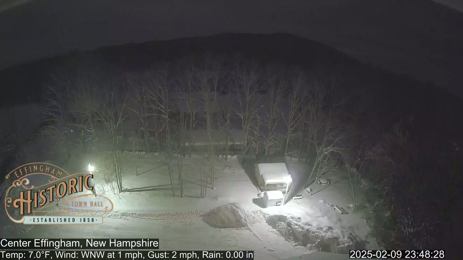A nighttime, snowy scene shows a snow-covered landscape with leafless trees, a small structure, and a lamppost under a dark sky.