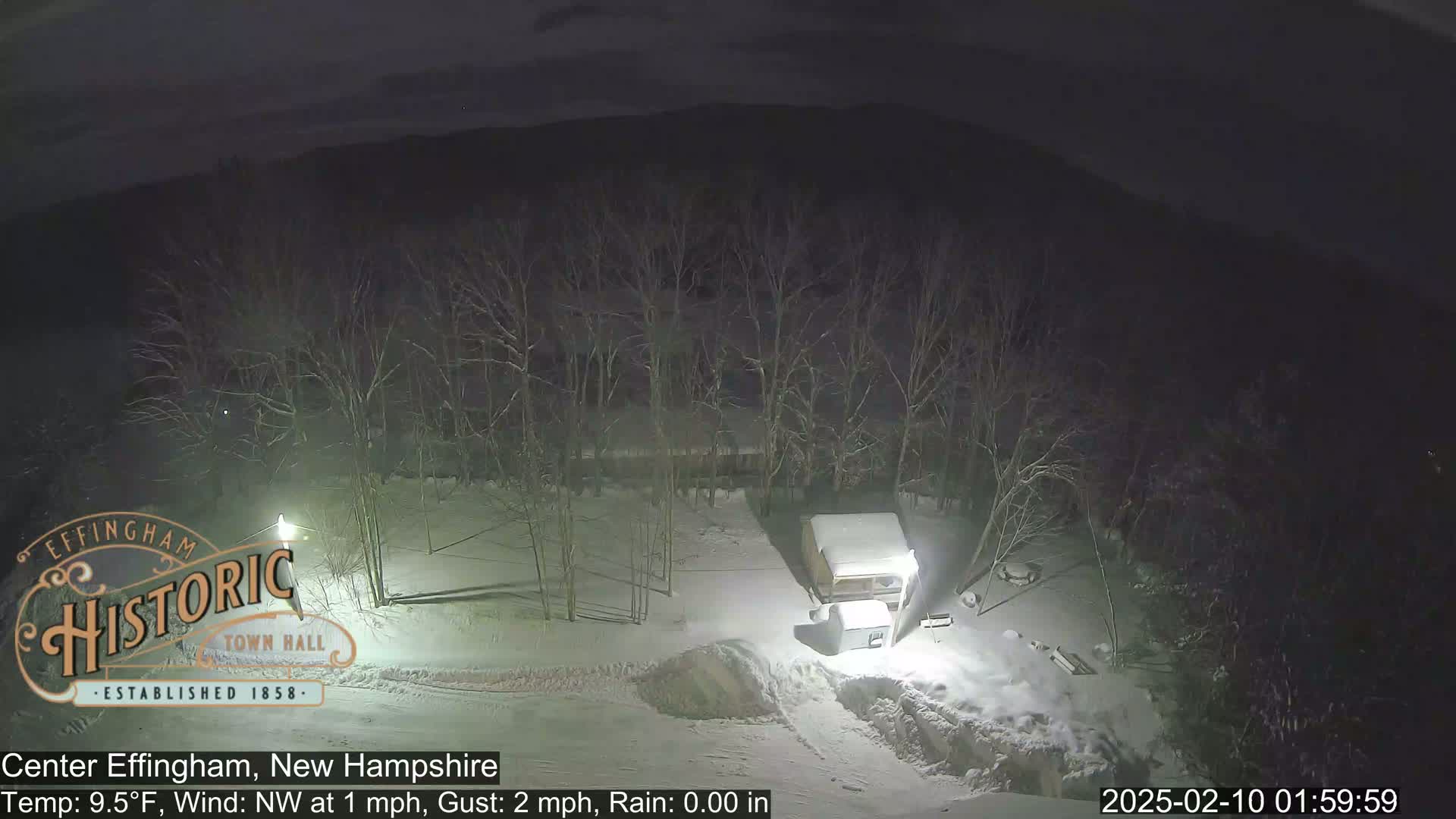 A snow-covered landscape at night, featuring bare trees and a small structure under a blanket of snow, with a light post illuminating the scene.