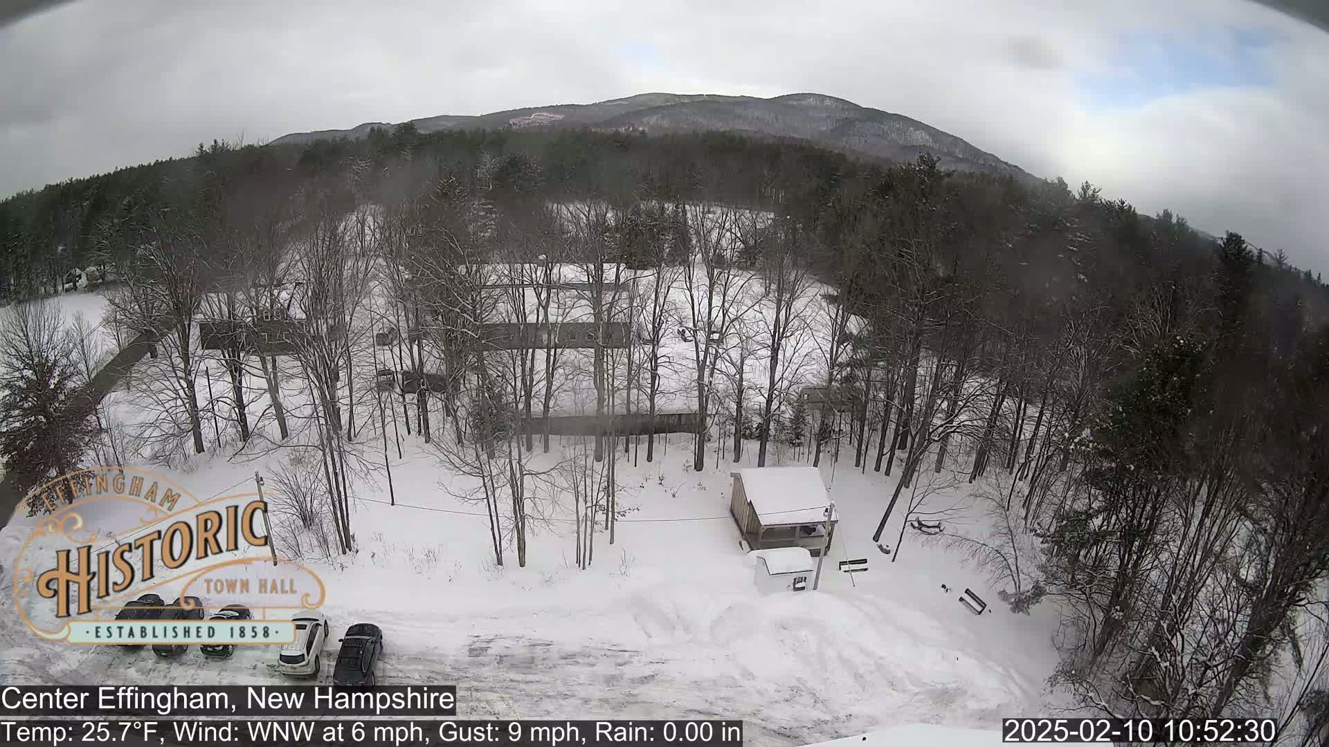 A snow-covered landscape with bare trees, a small building, and several vehicles parked in a lot, all under an overcast sky.