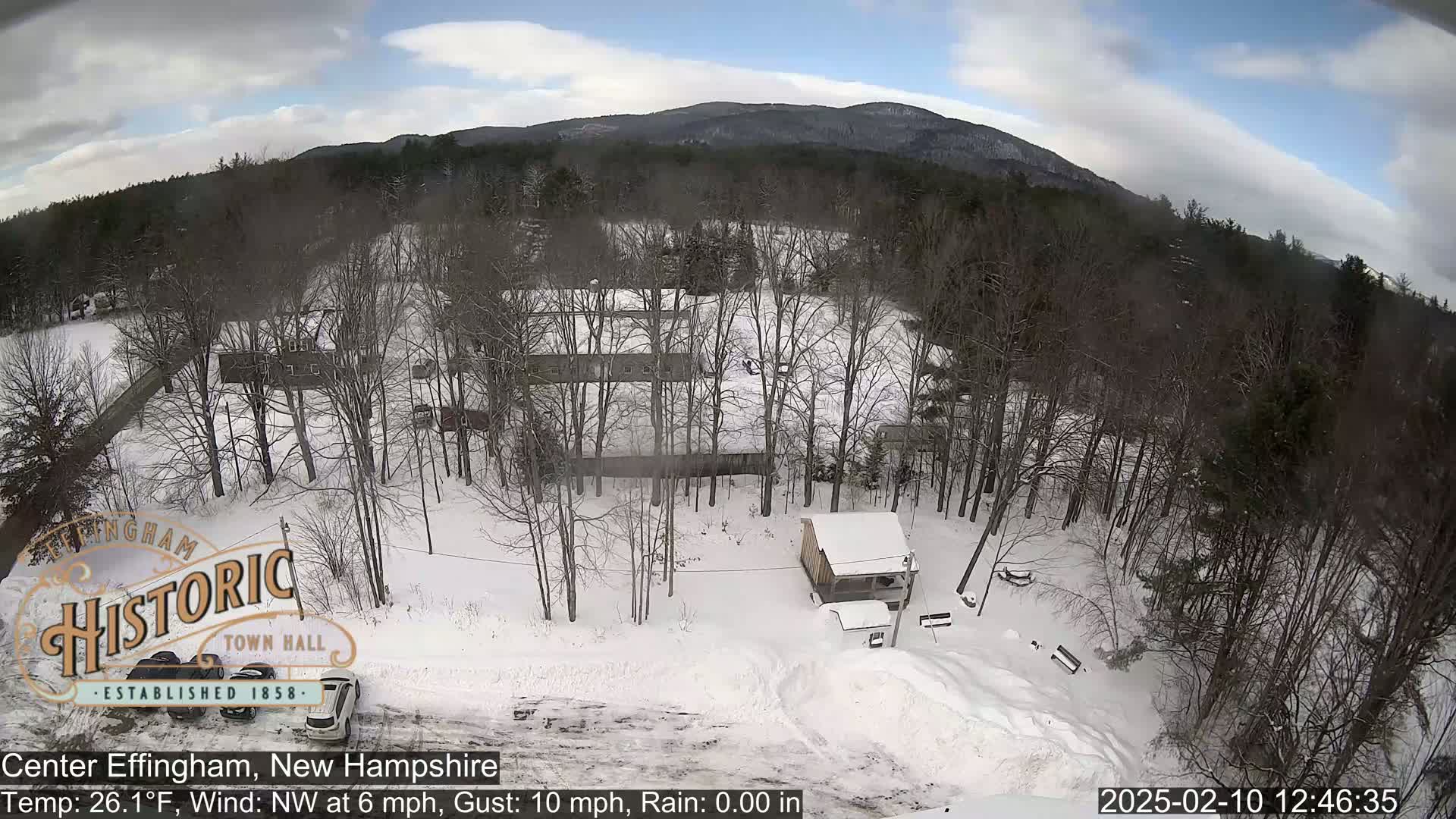 A snow-covered landscape with leafless trees, a small building, and mountains in the background under a partly cloudy sky.