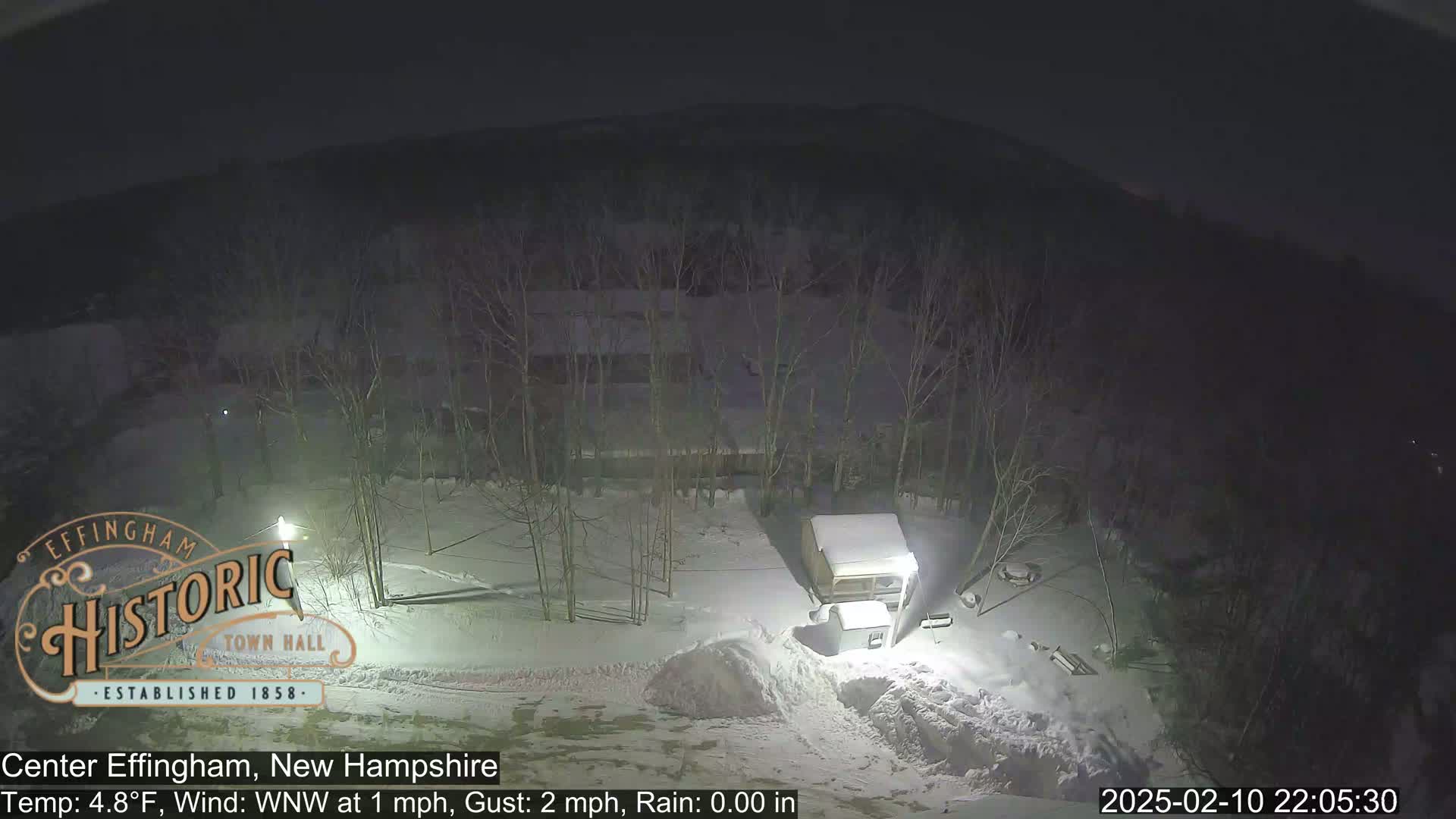 A nighttime, snowy scene shows a small building and snow-covered trees under a dark sky.