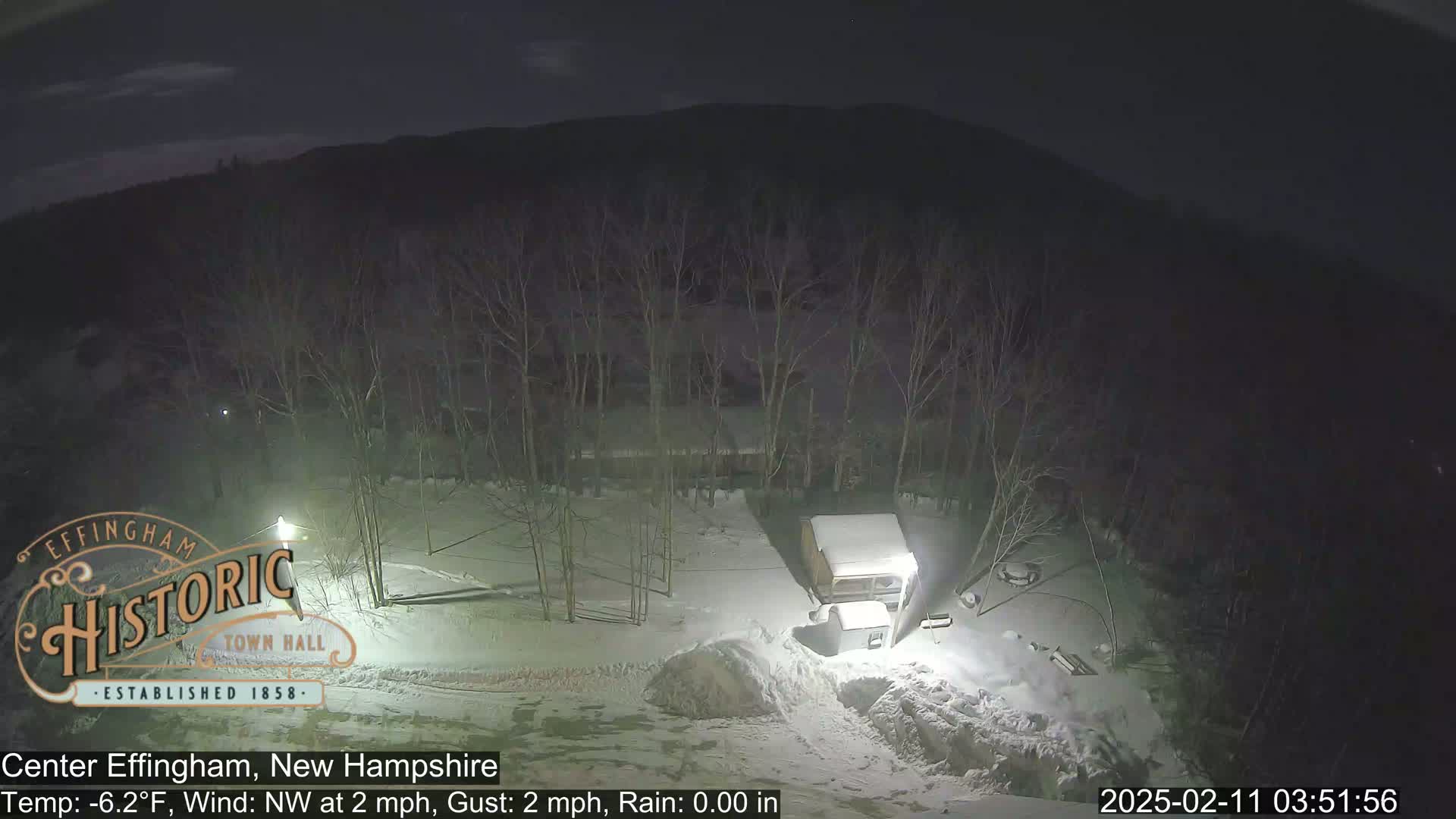 A nighttime, snow-covered scene shows a small building and trees under a dark sky with a northwest wind of 2 mph and a temperature of -6.2°F.