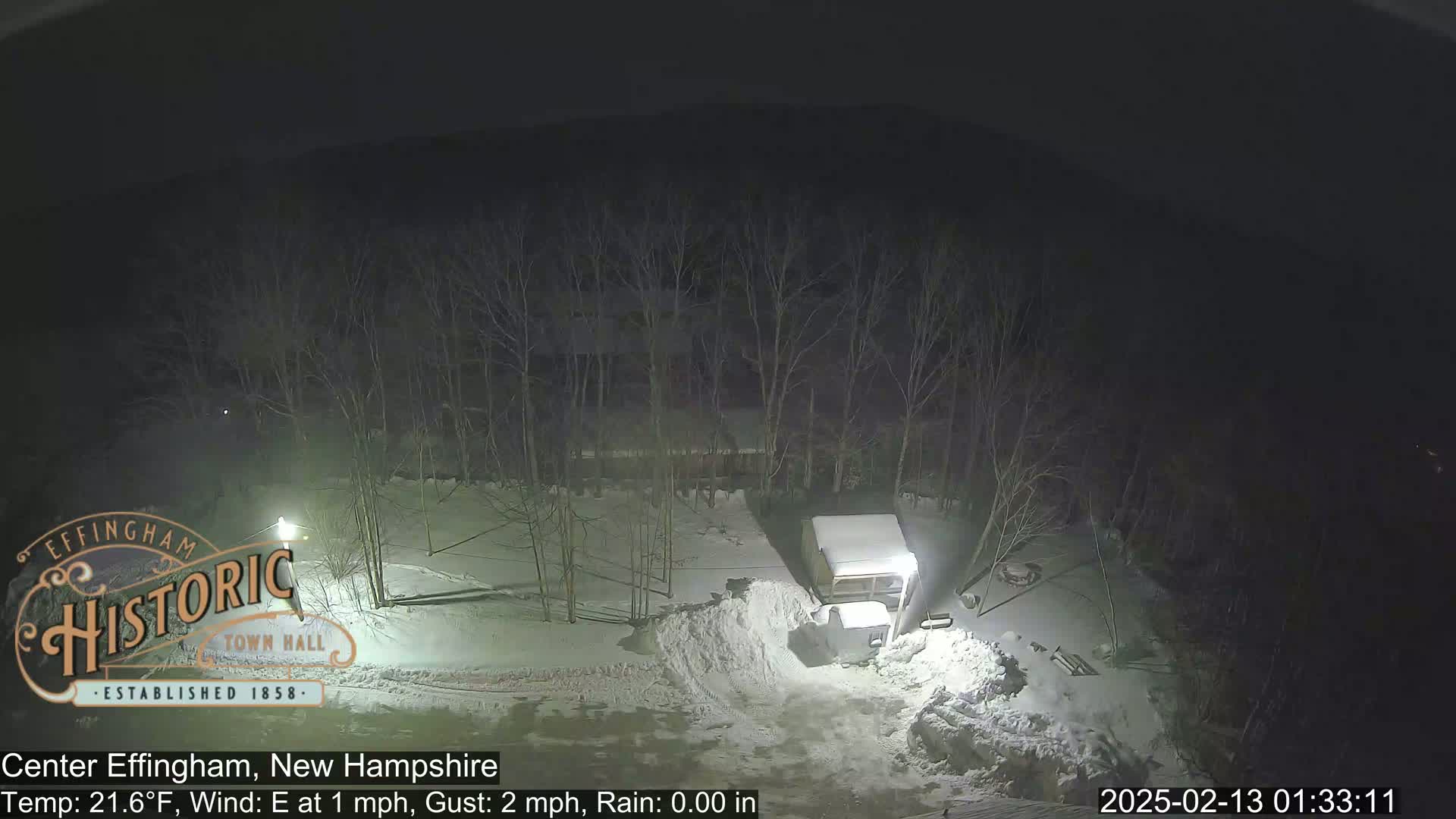 A nighttime scene of a snow-covered area with bare trees, a vehicle, and a pile of snow, under rainy conditions with an east wind.