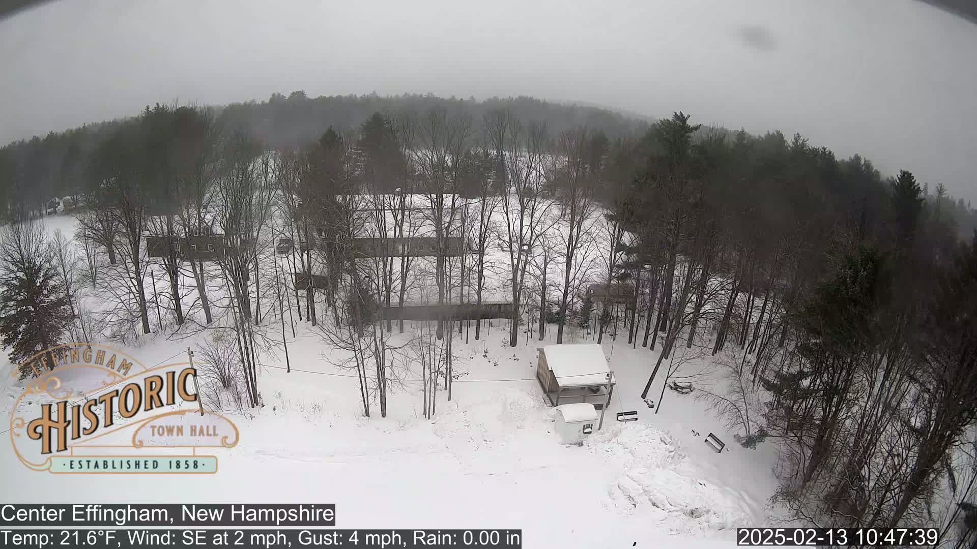 A snow-covered landscape with leafless trees and a small building is seen under an overcast sky.