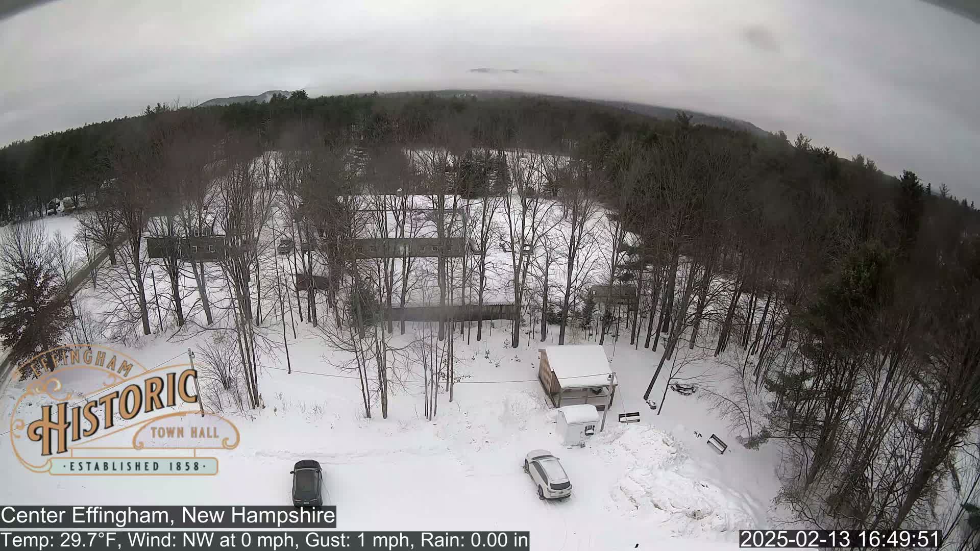 A snow-covered, wooded area with a few buildings and two cars parked in a lot under an overcast sky.