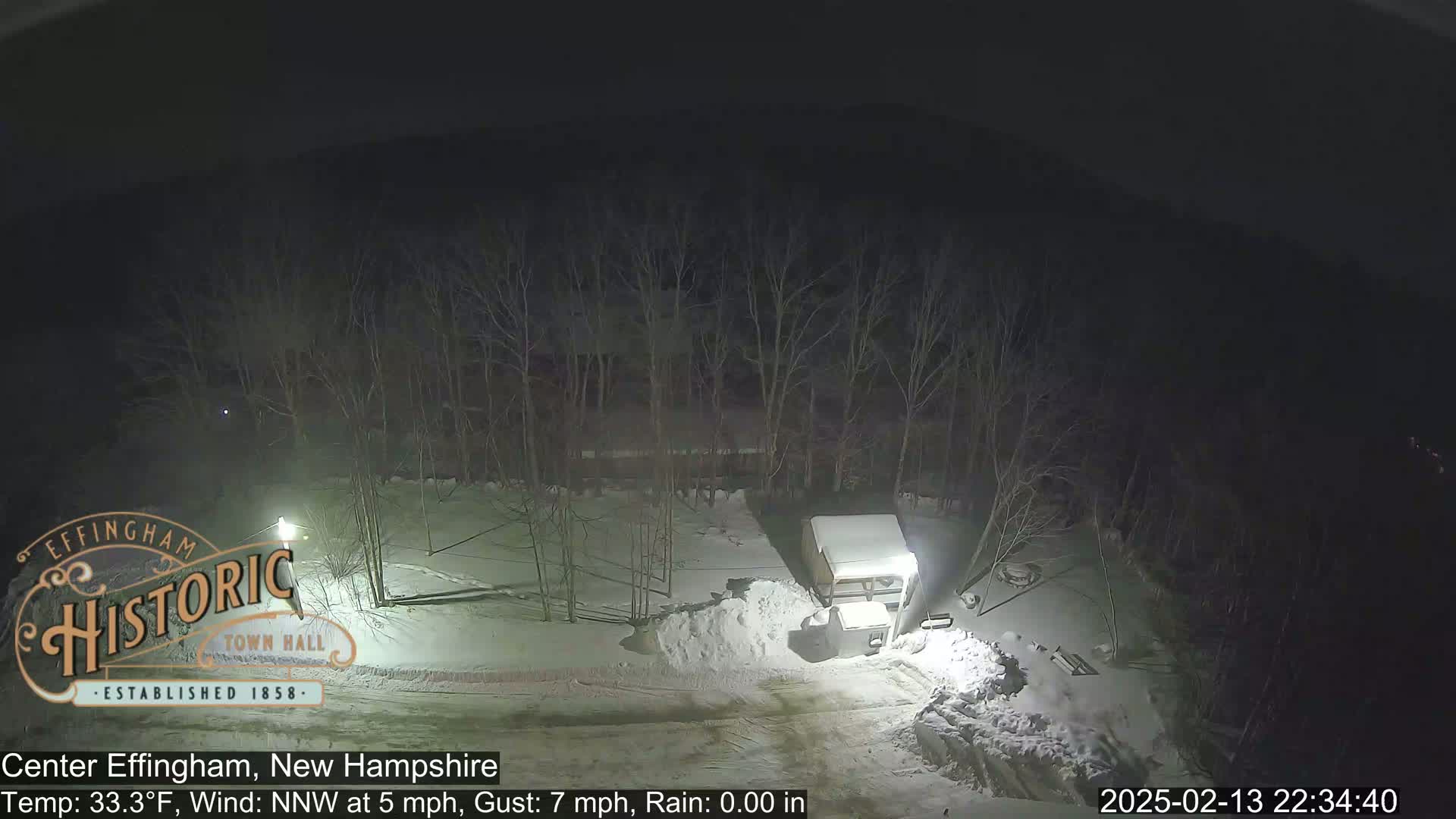 A snow-covered, nighttime scene shows a small, snow-covered vehicle parked near a pile of snow, surrounded by bare trees under a dark sky.