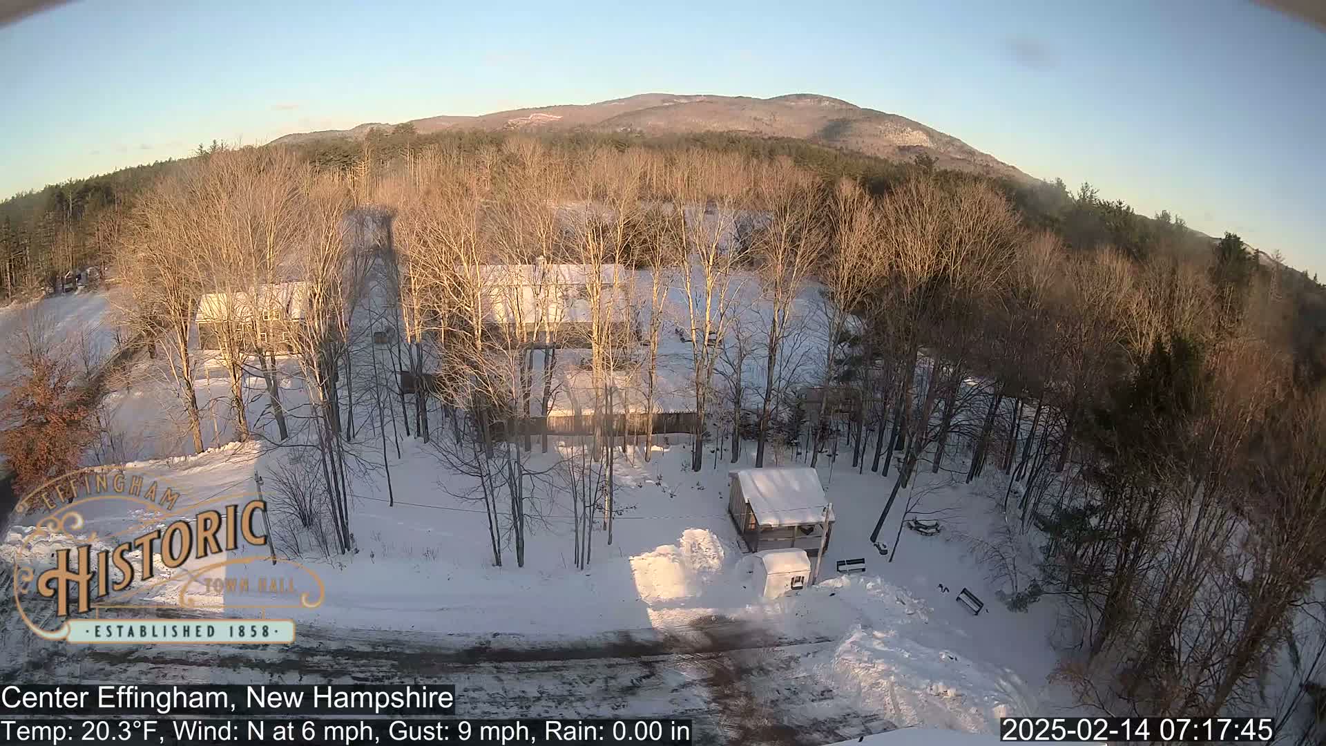 A snow-covered landscape with buildings, bare trees, and a mountain in the background under a clear, sunny sky.