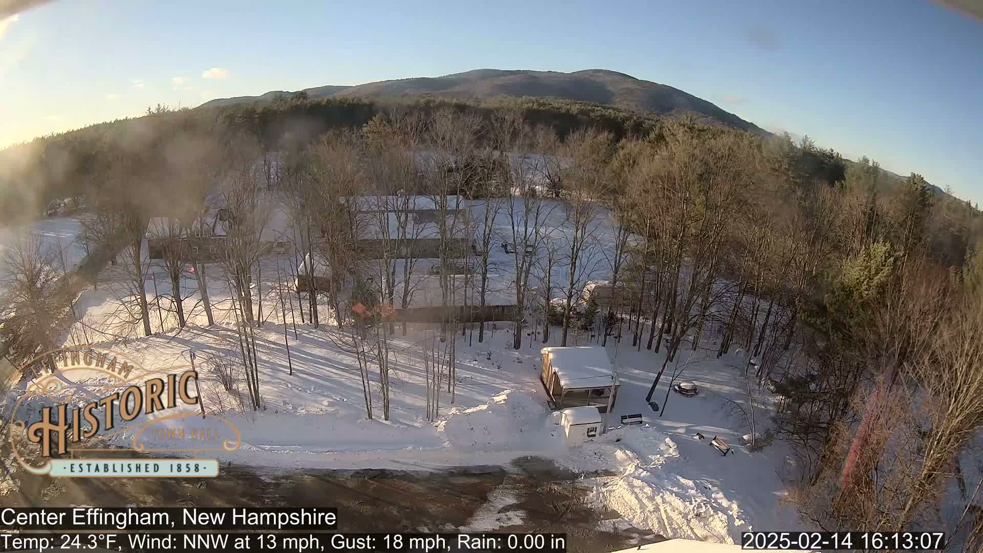 A snow-covered landscape with several buildings and bare trees, under a sunny sky, shows a view of a town in a mountainous region.