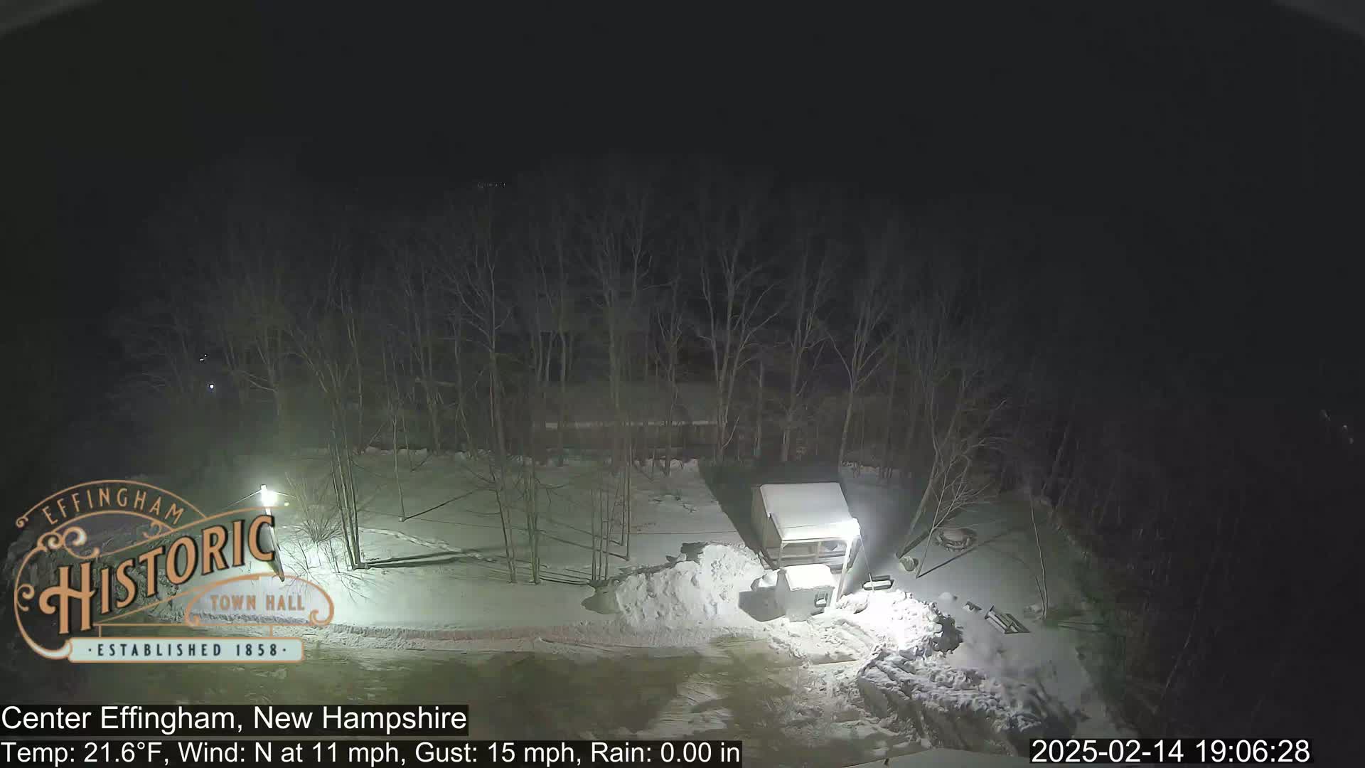 A snow-covered nighttime scene shows a camper and a pile of snow near bare trees under a dark sky.