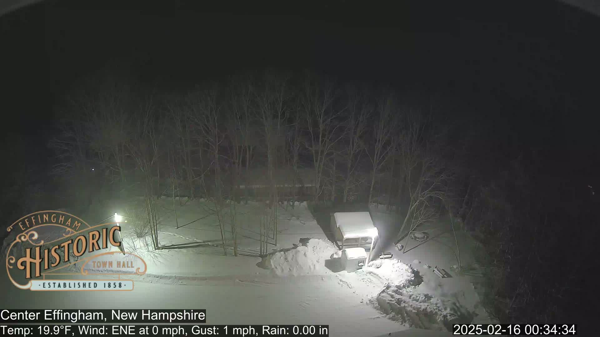 A snow-covered scene at night shows a structure and a pile of snow near bare trees under a dark sky.