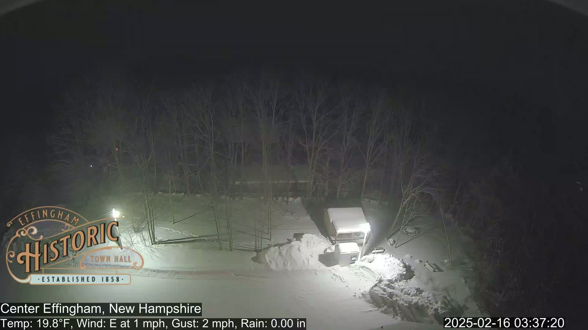 A nighttime, snowy scene shows a snow-covered vehicle parked near a snowdrift, with bare trees in the background under a dark sky.