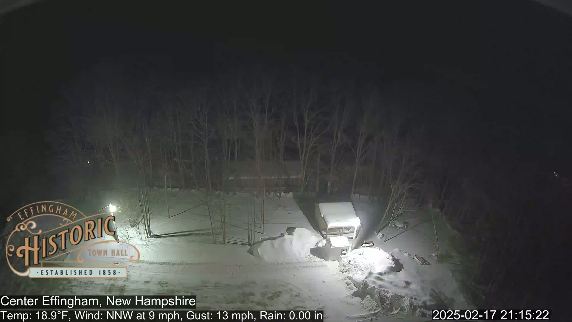 A snow-covered, nighttime scene shows a small structure and a pile of snow near bare trees under a dark sky with light rain.