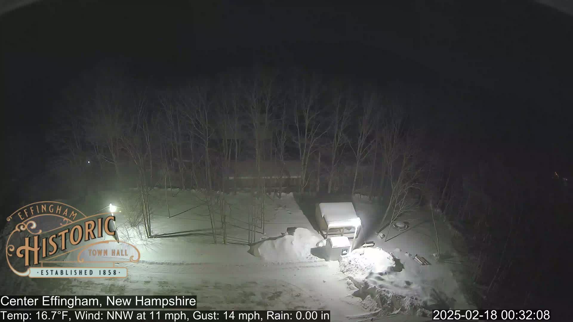 A snow-covered scene at night shows a camper, a lamp post, and bare trees under a dark sky.