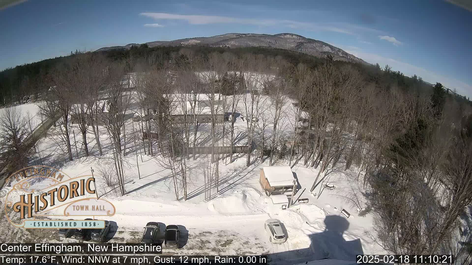 A snow-covered parking lot and surrounding wooded area with a few buildings are visible under a sunny sky.