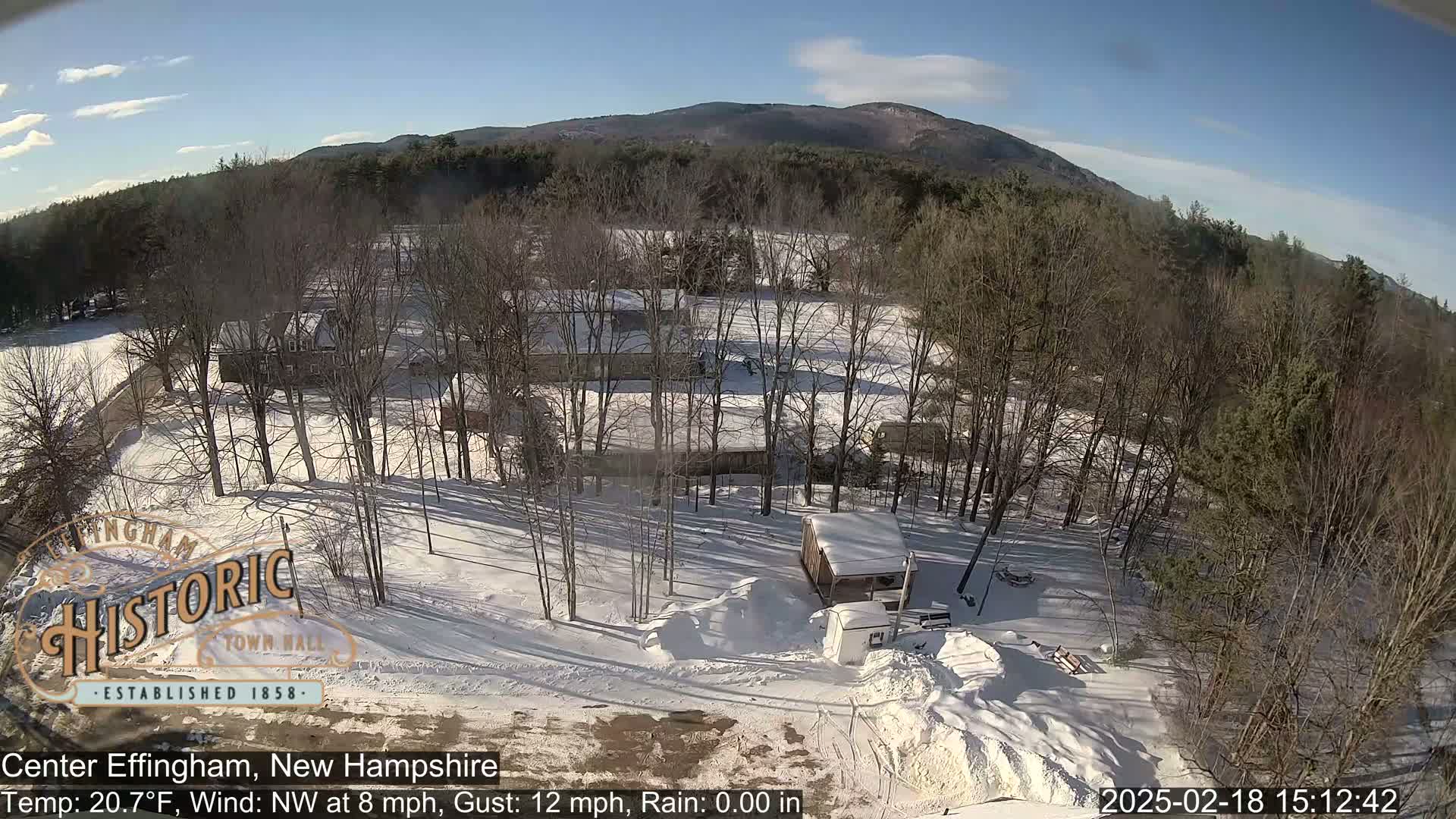 A snow-covered landscape with bare trees, a few buildings, and a mountain in the background under a mostly sunny sky.