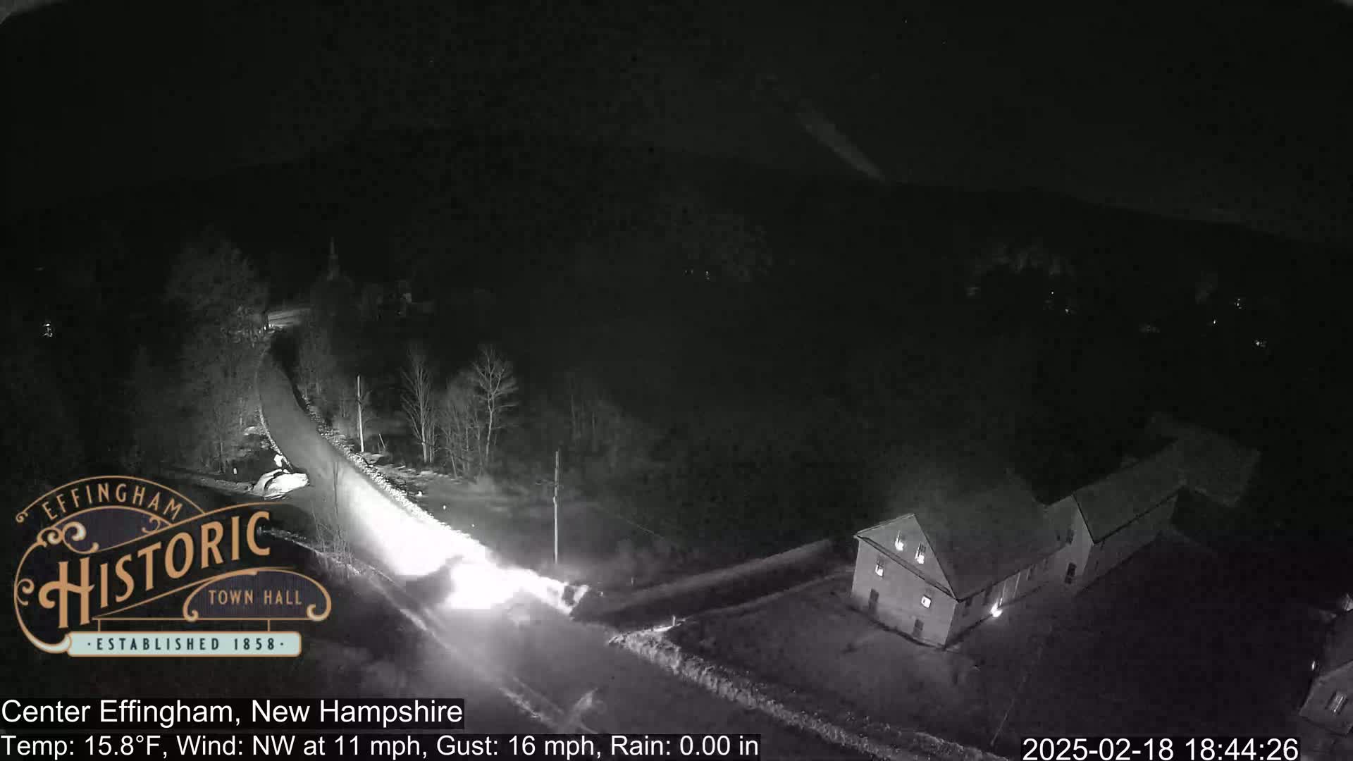 A nighttime, snow-covered road scene shows a vehicle's headlights illuminating the road and a building in the lower right, with a northwest wind at 11 mph and gusts up to 16 mph.