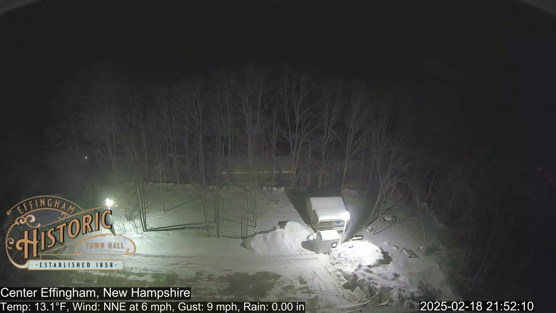 A snow-covered, nighttime scene shows a camper parked near a snow pile next to bare trees under a dark sky.