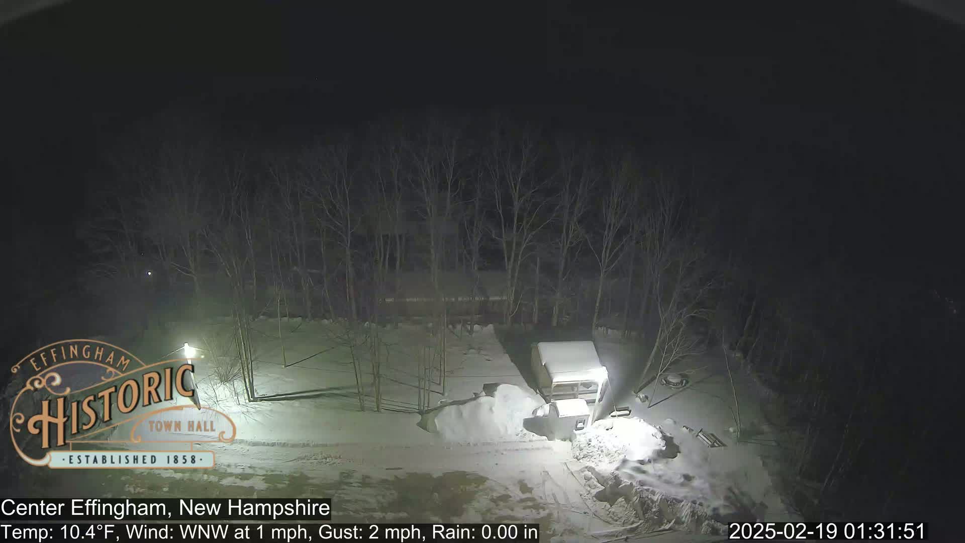 A nighttime, snow-covered scene shows a camper and a pile of snow near bare trees under a dark sky.