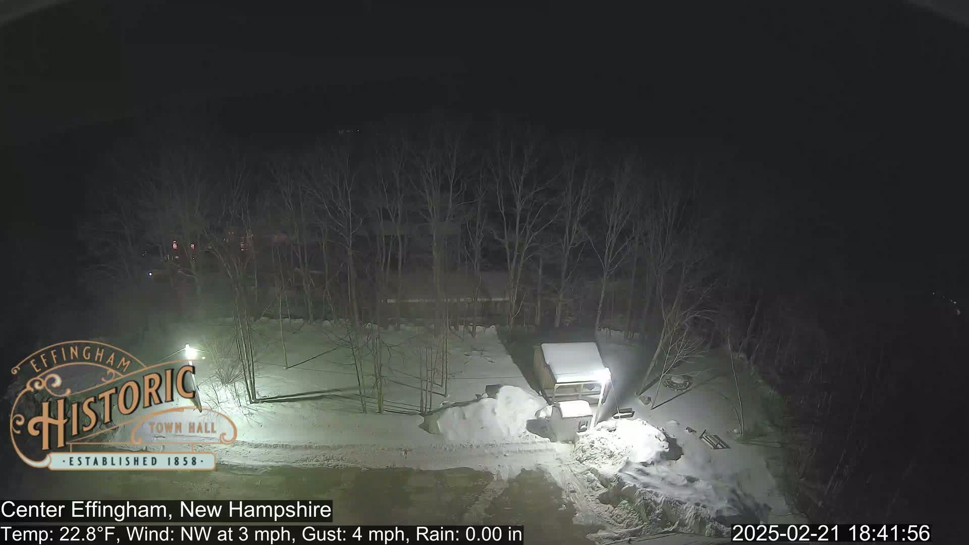 A nighttime, snow-covered scene shows bare trees surrounding a small building and a pile of snow next to a road under a dark sky.