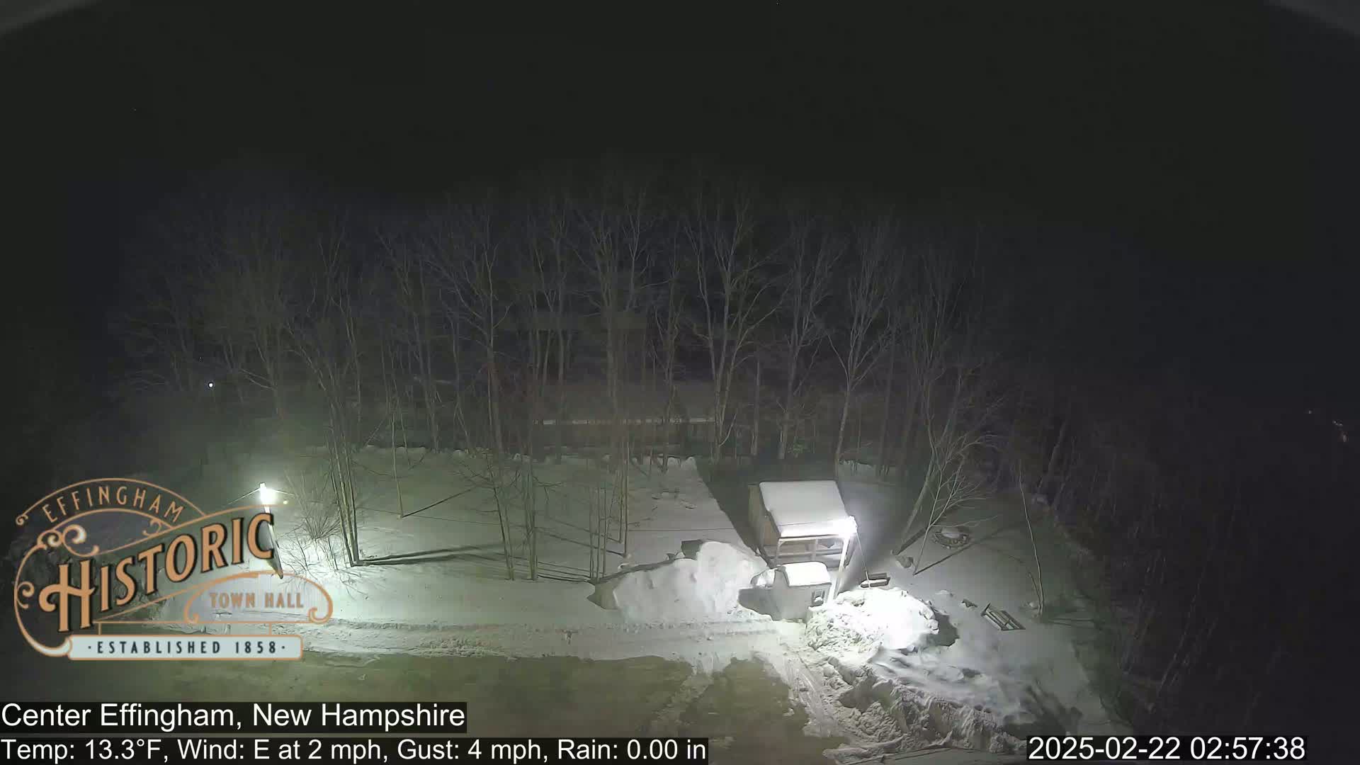 A snow-covered area at night, illuminated by a light post, with bare trees in the background, under a dark sky with a temperature of 13.3°F, an easterly wind of 2 mph with gusts up to 4 mph, and no rain.