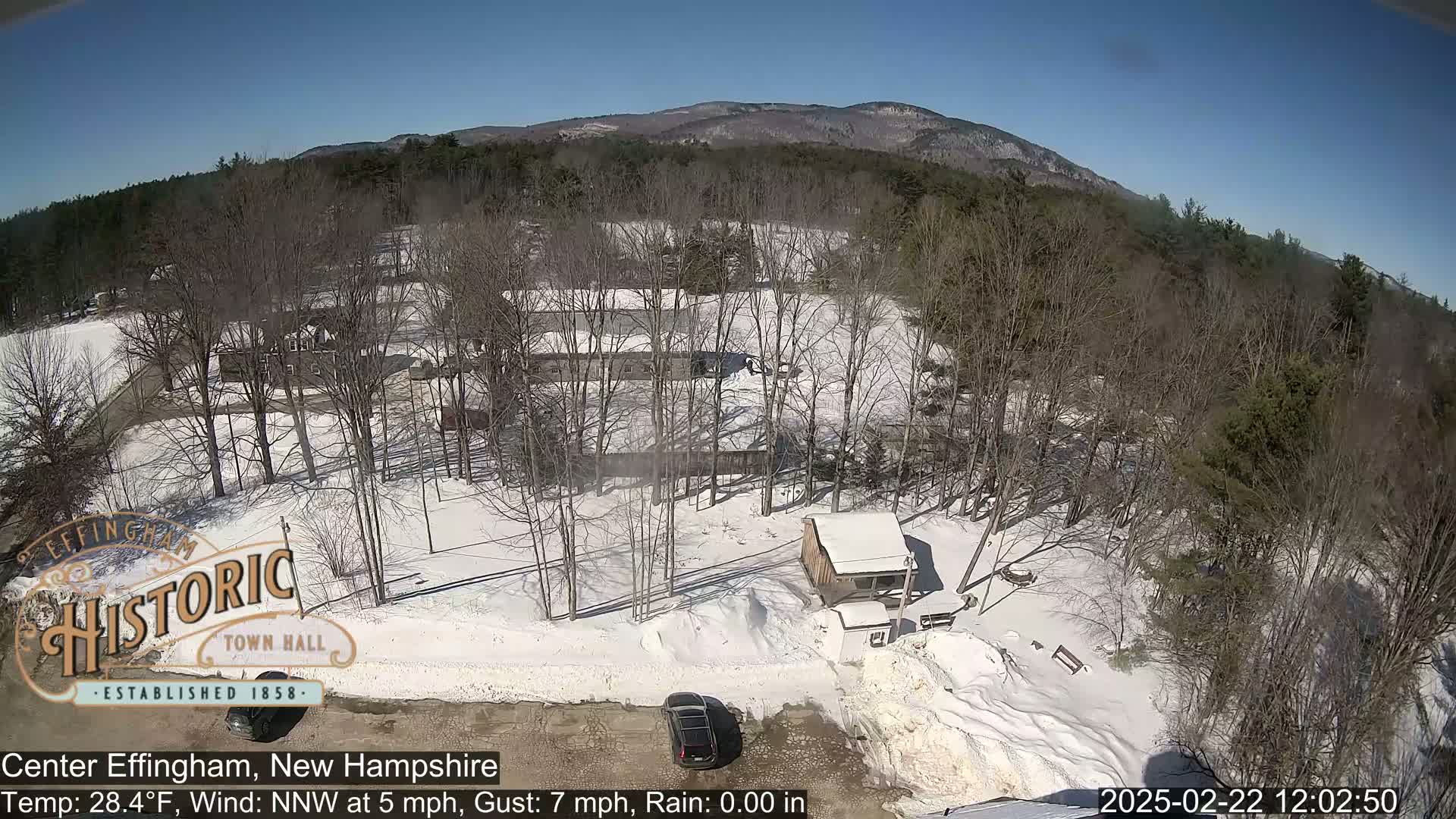 A snow-covered landscape with leafless trees, a small building, and a few cars is visible under a clear blue sky.