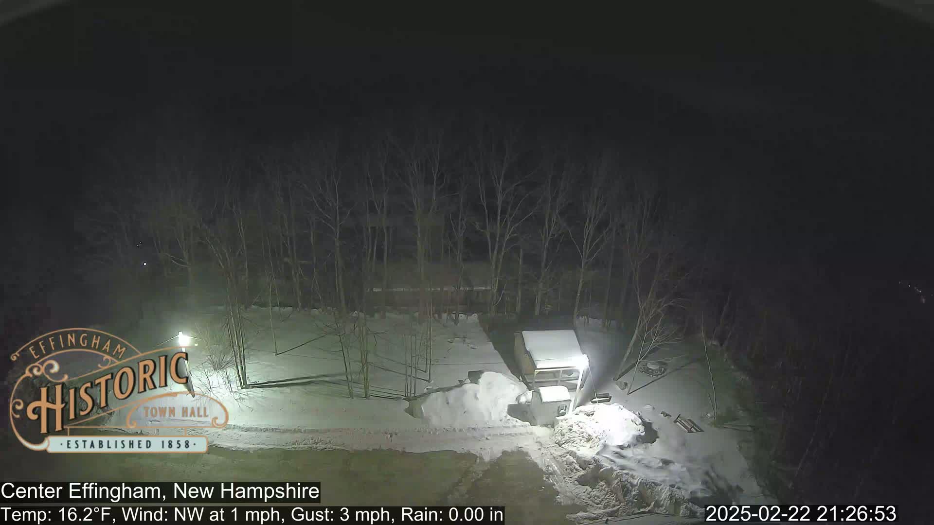 A snow-covered nighttime scene shows bare trees surrounding a building and a pile of snow, with a light post illuminating a portion of the area.