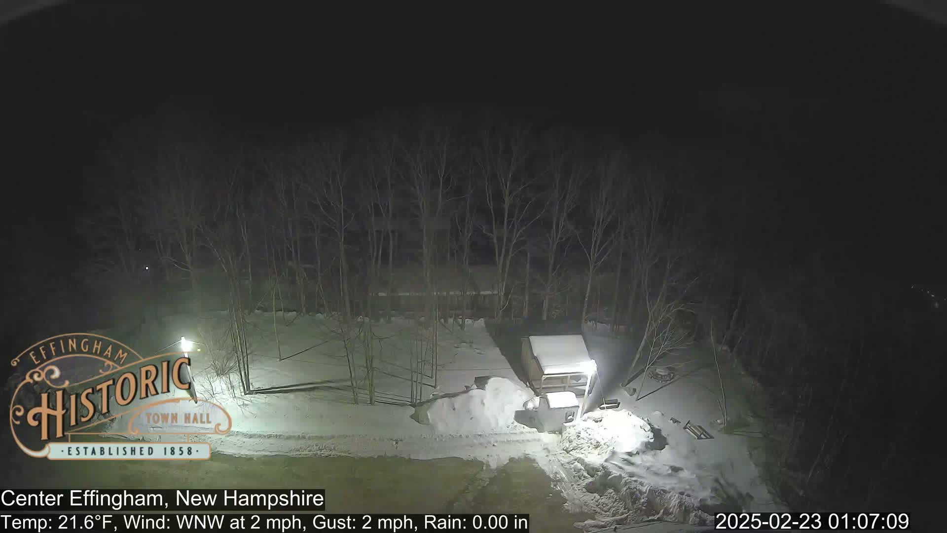 A nighttime scene of a snow-covered area with bare trees, a small structure, and a vehicle, under a dark sky with a light wind.