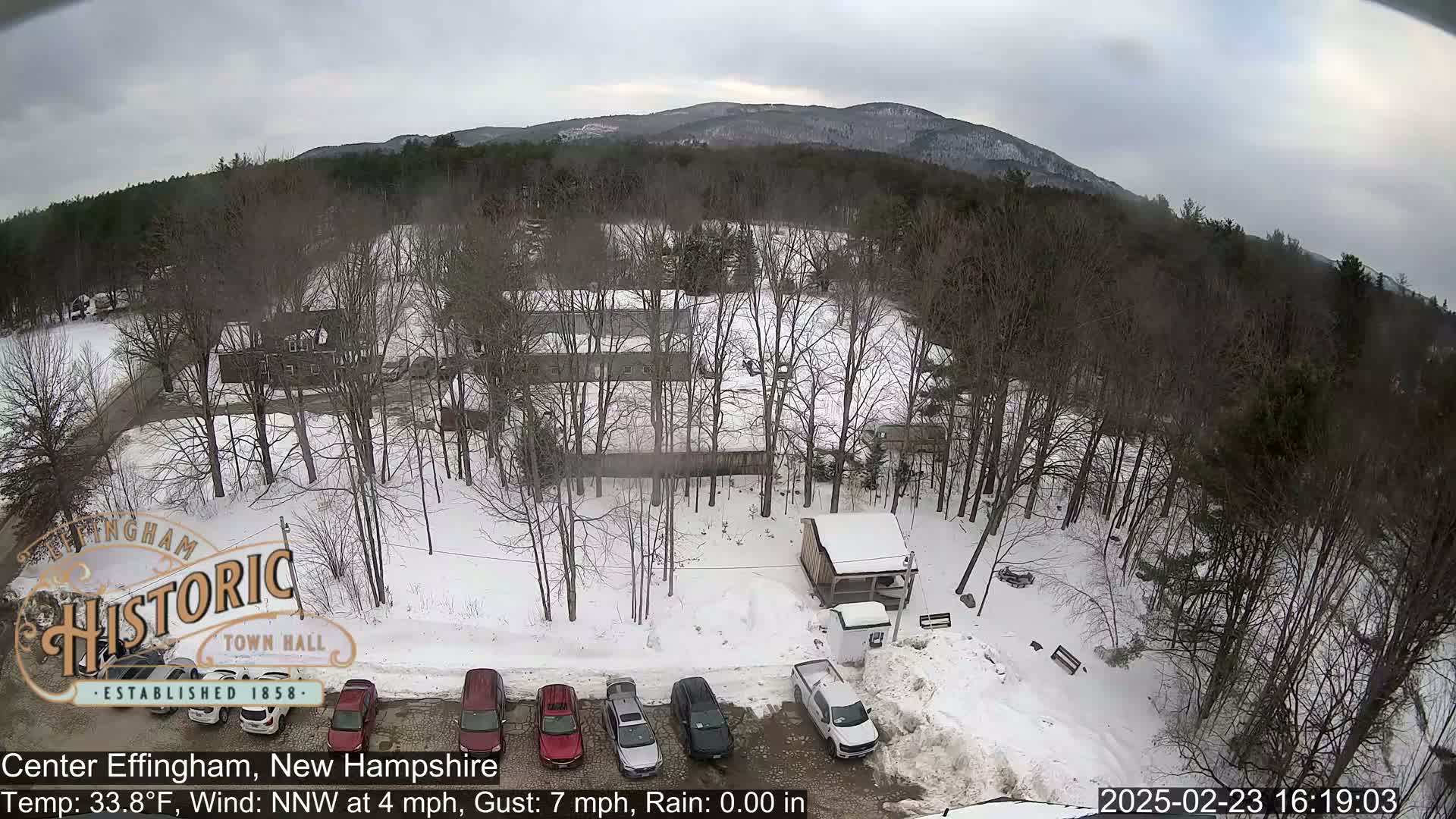 A snow-covered parking lot with several cars, a small building, and leafless trees, all set against a backdrop of a distant mountain range under an overcast sky.