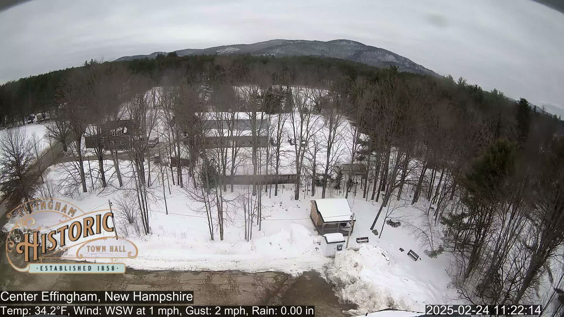A snow-covered landscape with leafless trees, a small building, and a road, under a cloudy sky.