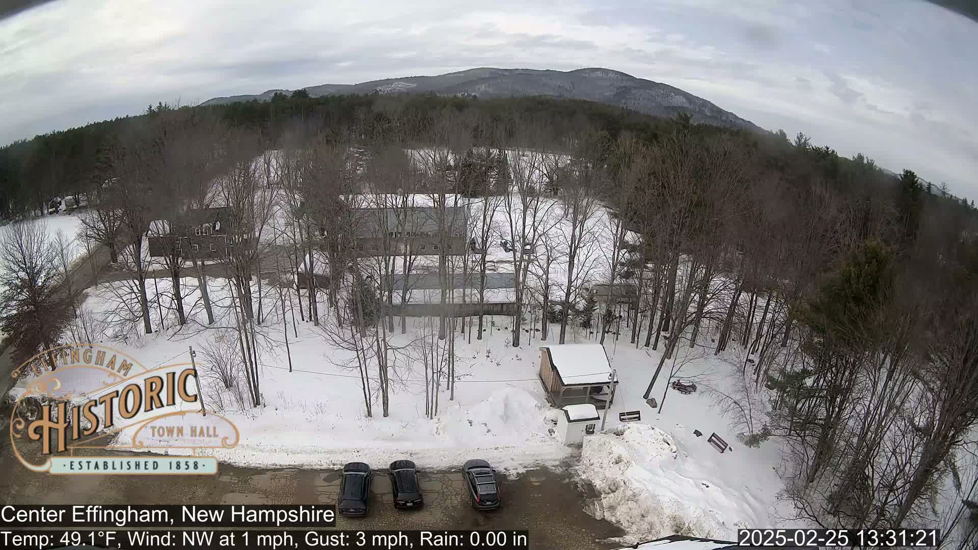 A snow-covered parking lot with several cars, a small building, and bare trees in front of a snow-covered field, all set against a backdrop of distant mountains under an overcast sky.