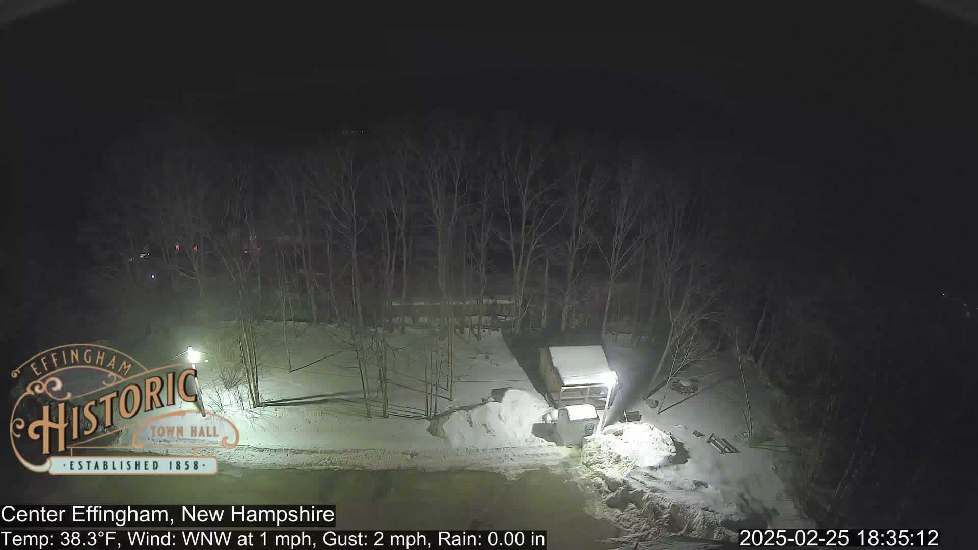 A snow-covered nighttime scene shows a small building and piles of snow near bare trees under a dark sky.