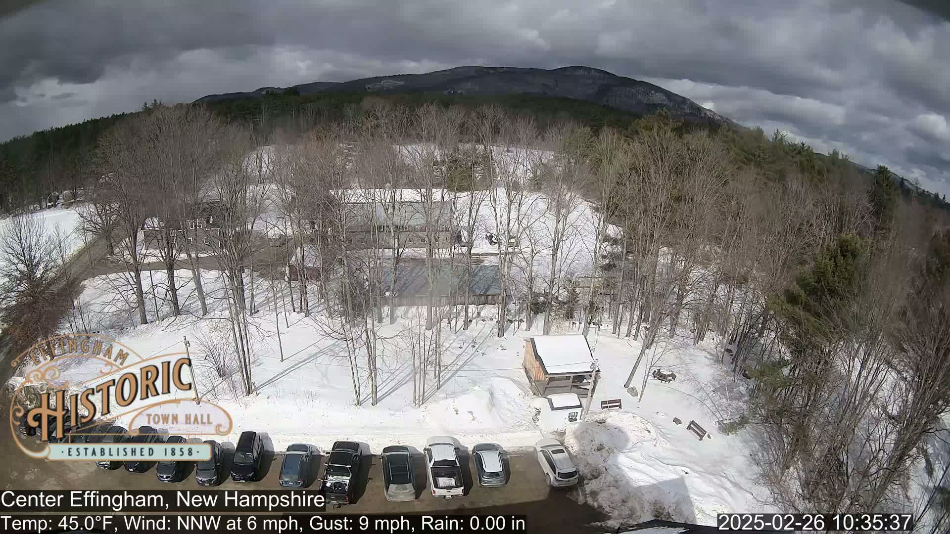 A snow-covered parking lot with several cars and buildings is nestled among leafless trees, with mountains visible in the background under a partly cloudy sky.