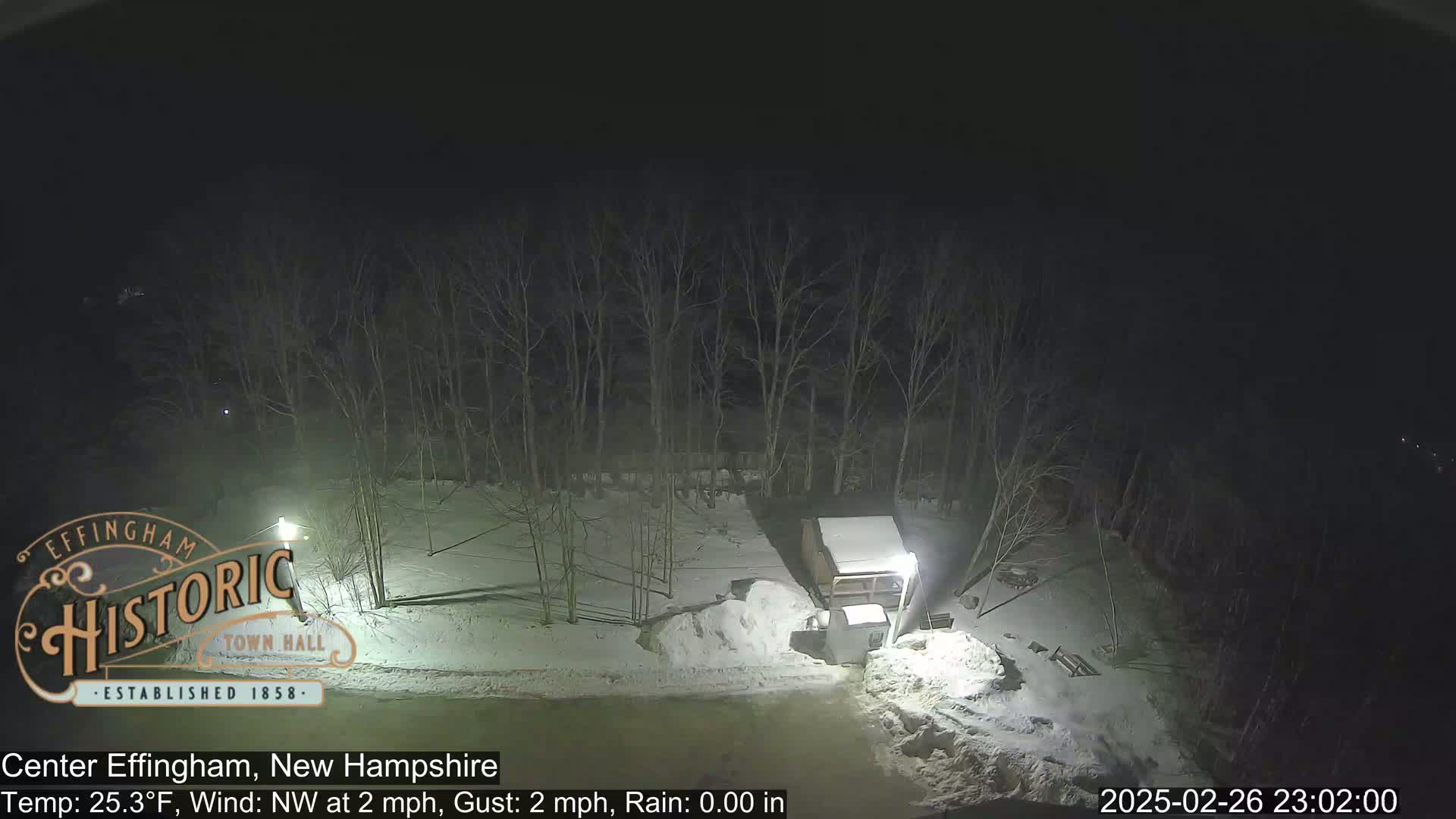 A nighttime, snow-covered scene shows a small shed and bare trees under a dark sky.