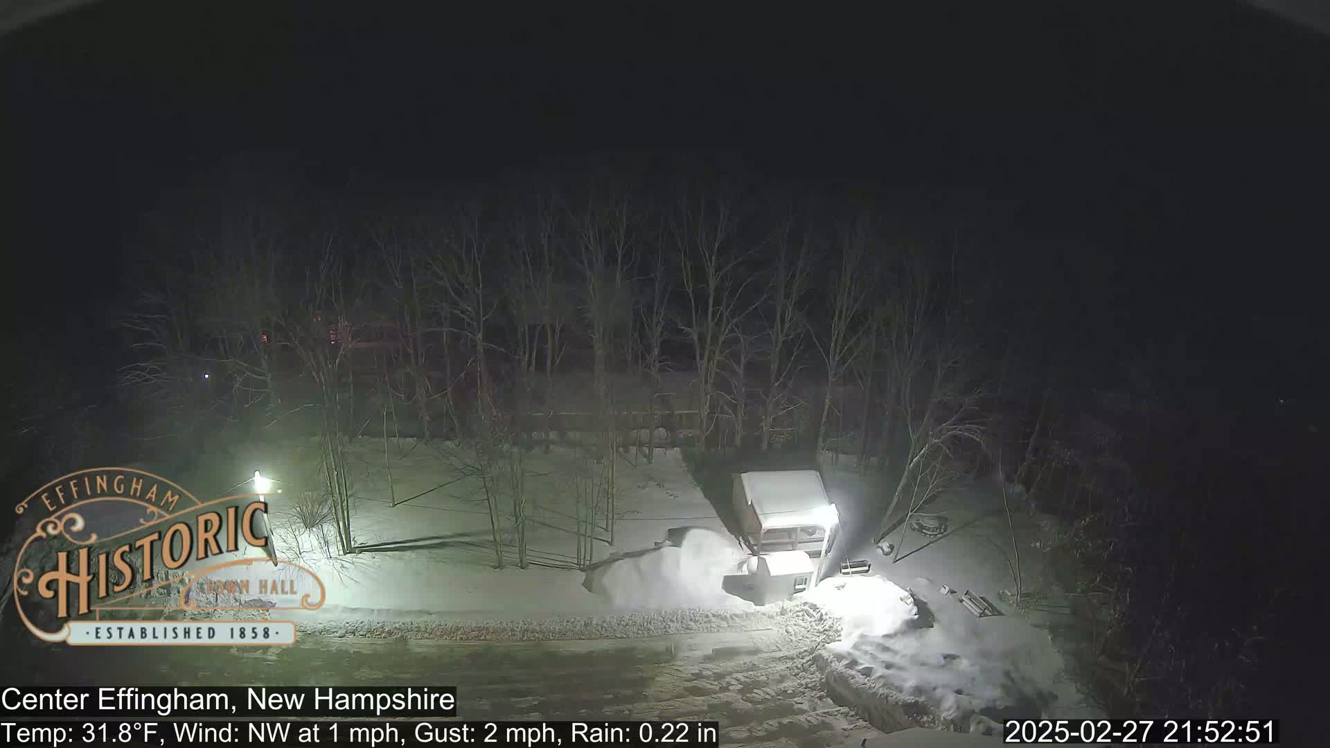 A nighttime scene shows a snow-covered area with bare trees and a structure partially obscured by snow, under conditions of light rain and a northwest wind.