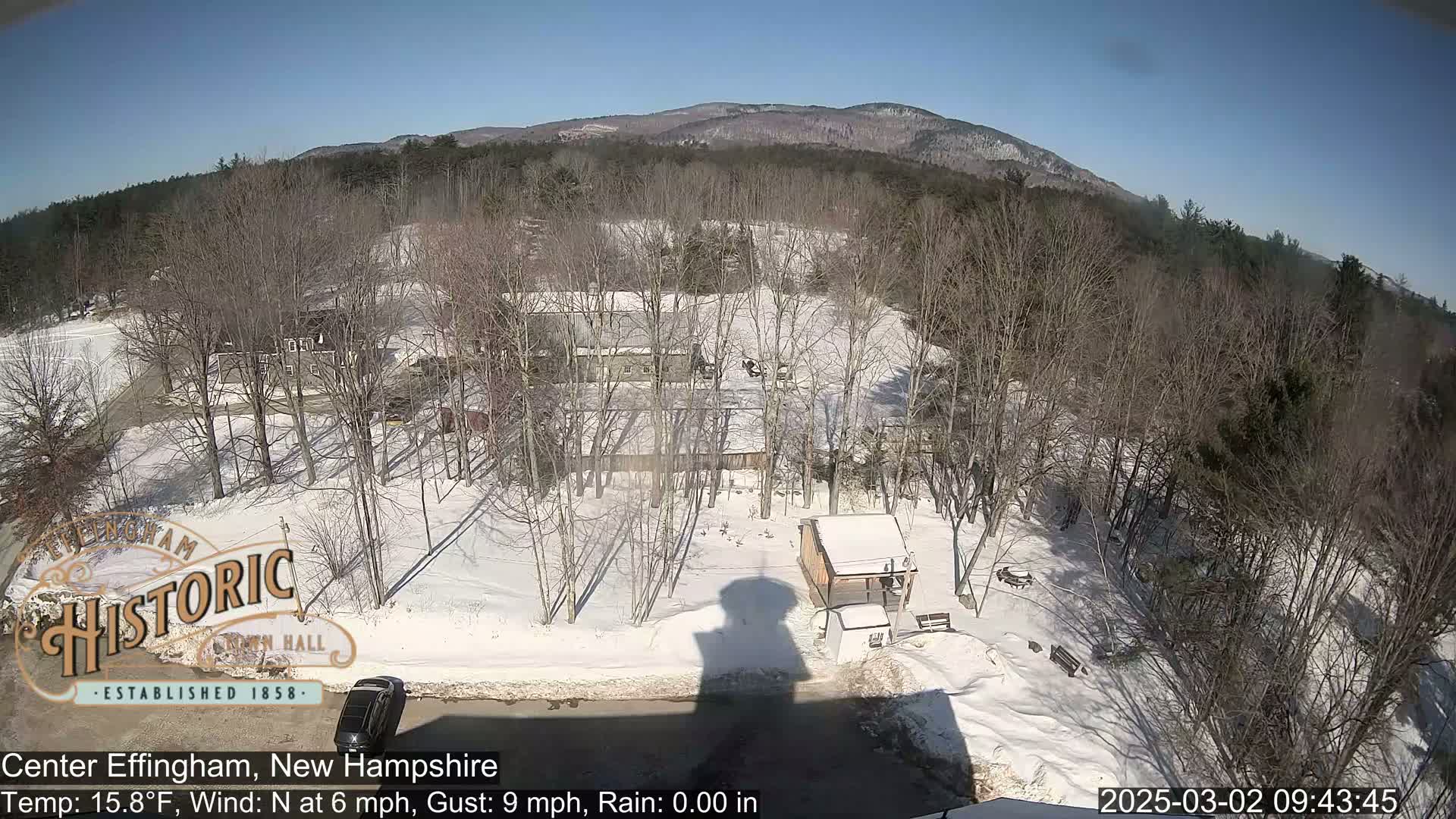 A snow-covered landscape with bare trees, a few buildings, and a mountain in the background under a sunny sky.