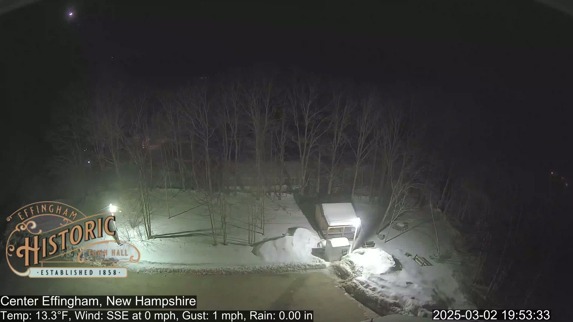 A nighttime view of a snow-covered area with bare trees and a small structure, under a clear sky.