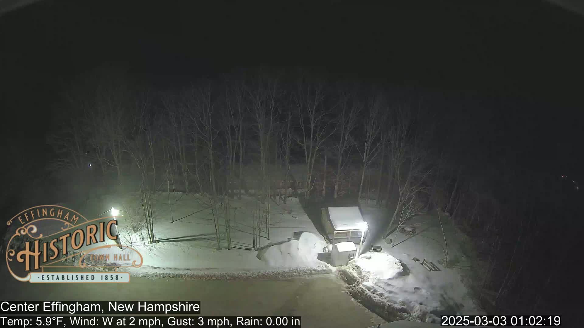 A nighttime view of a snow-covered area with bare trees, a small structure, and some snow piles, under a dark sky.