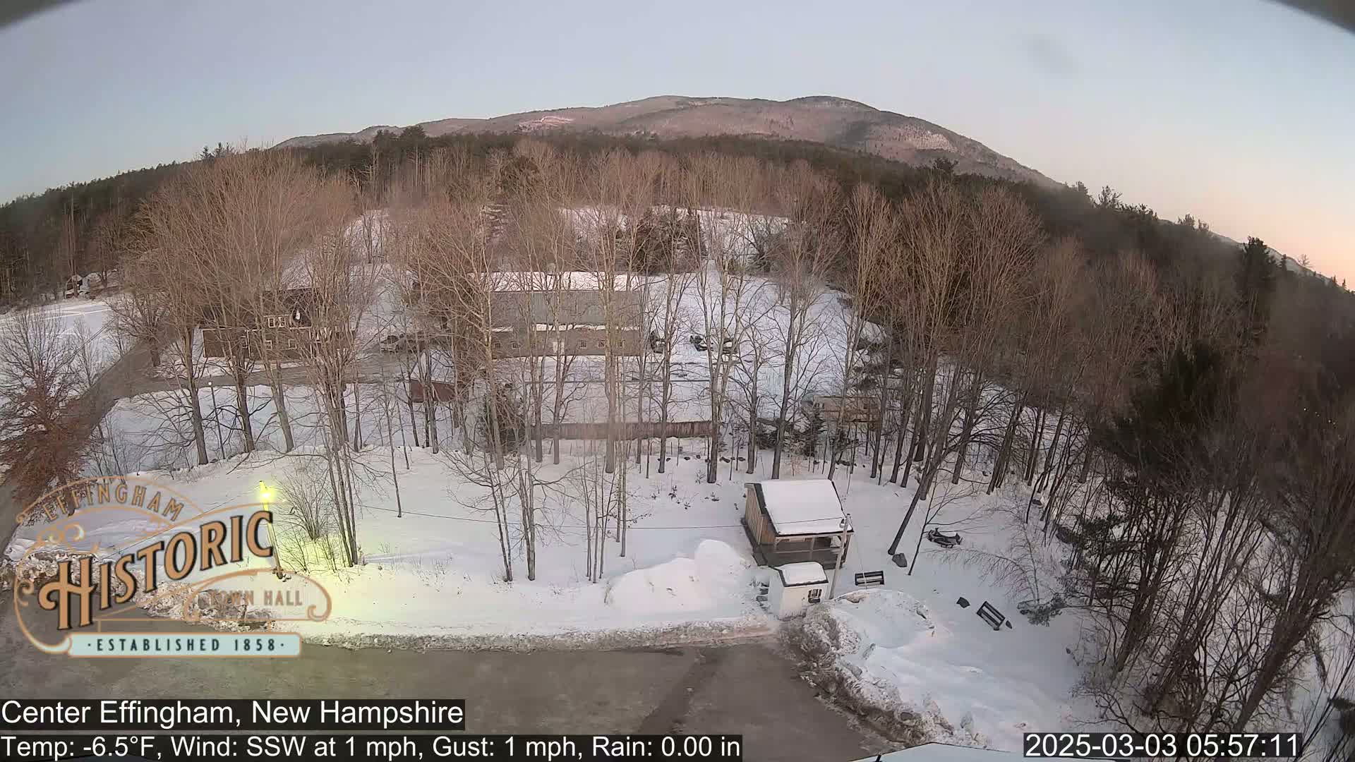 A snow-covered landscape with bare trees, a small building, and a mountain in the background under a twilight sky.