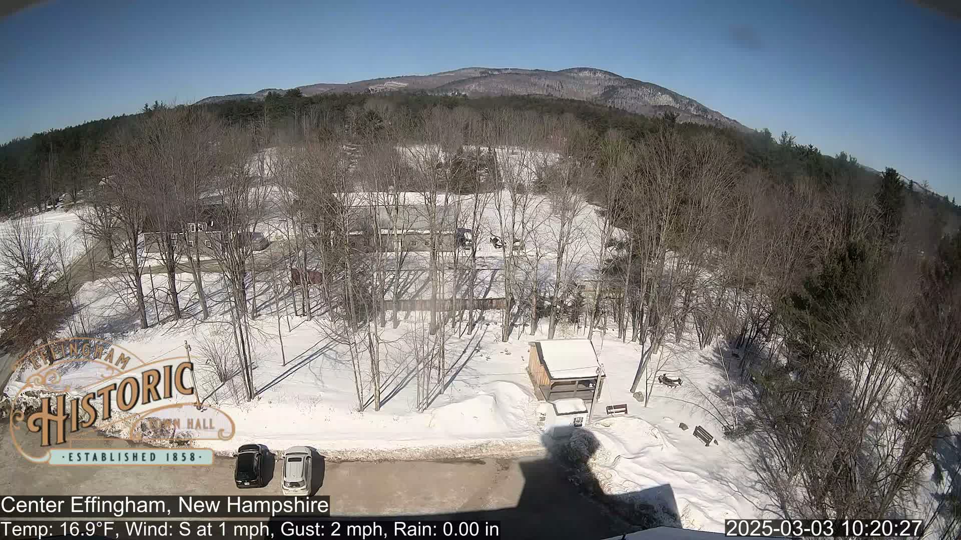 A snow-covered landscape with bare trees, a small building, and parked cars is visible under a sunny sky, with mountains in the background.