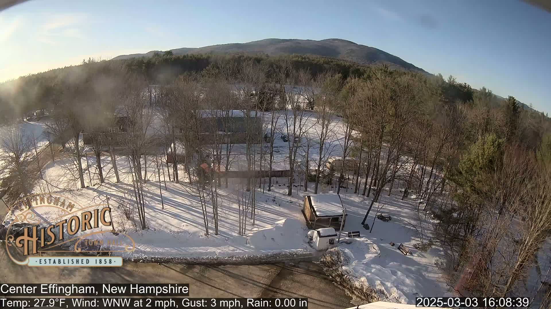 A snow-covered landscape with bare trees and buildings, under a sunny sky, with mountains in the background.