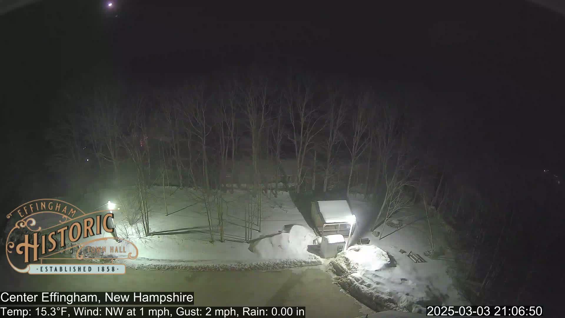 A nighttime scene of a snow-covered area with bare trees, a small building, and a pile of snow, under a clear sky.