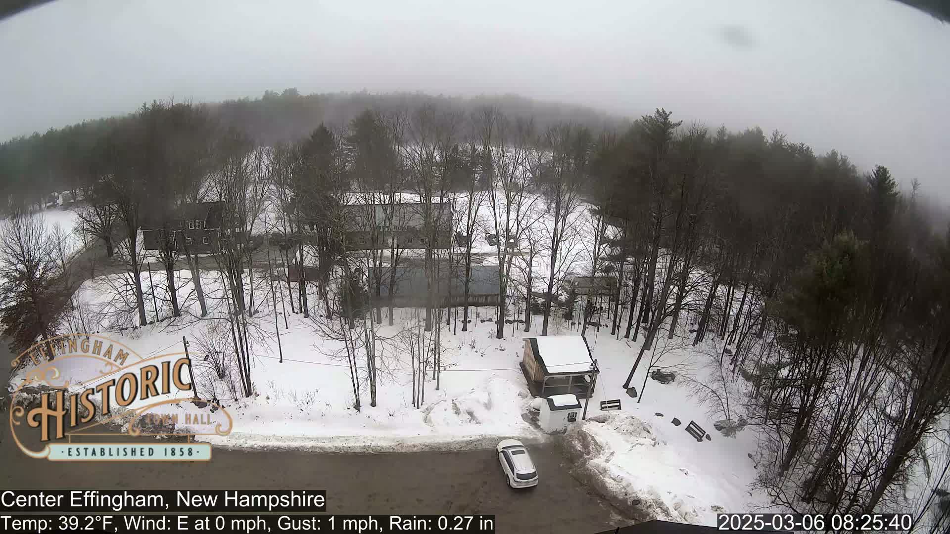 A snowy, overcast scene shows a parking lot with a car, several buildings, and a wooded area.