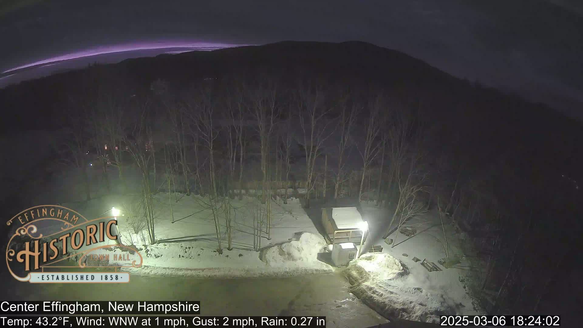 A nighttime scene of a snow-covered area with bare trees, a small structure, and a faint purplish glow on the horizon under rainy conditions.