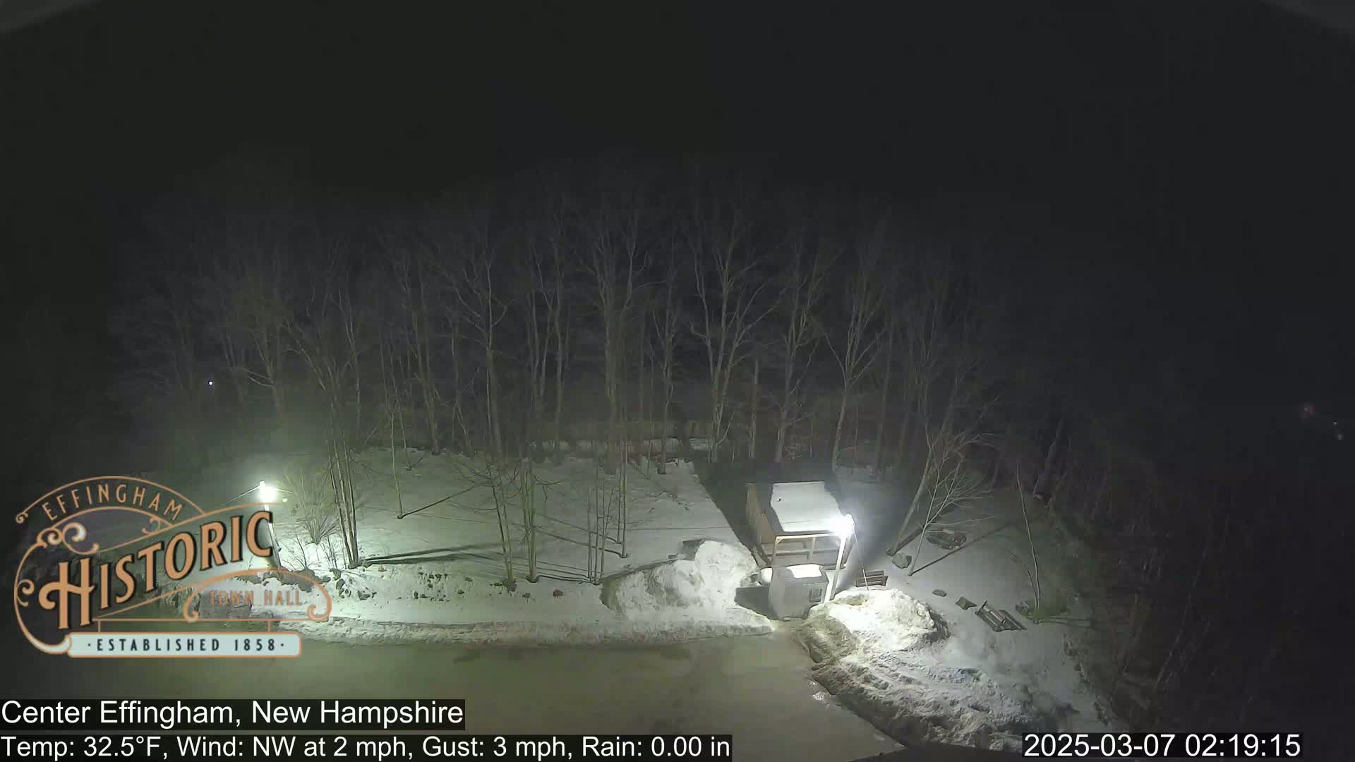 A nighttime view of a snow-covered area with leafless trees and a small building under partly cloudy skies.