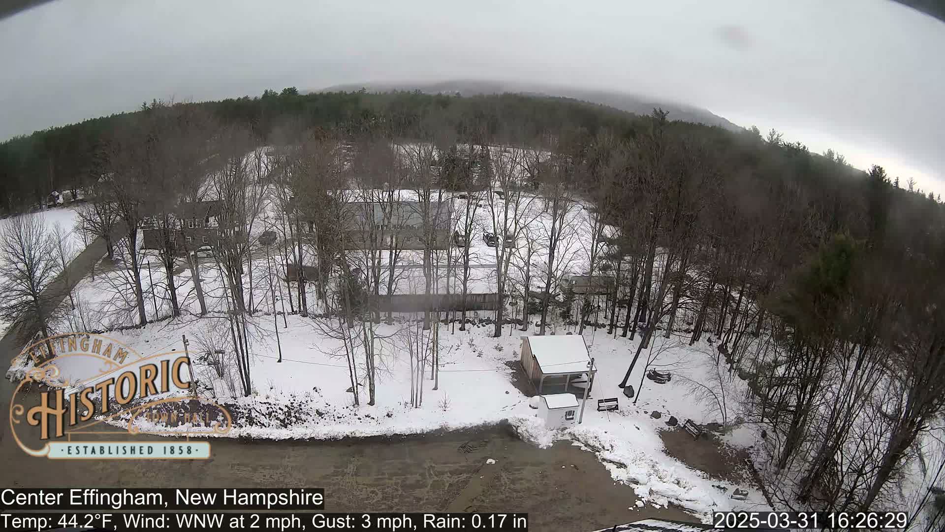A snow-covered landscape with bare trees and several buildings, under an overcast sky with light rain.