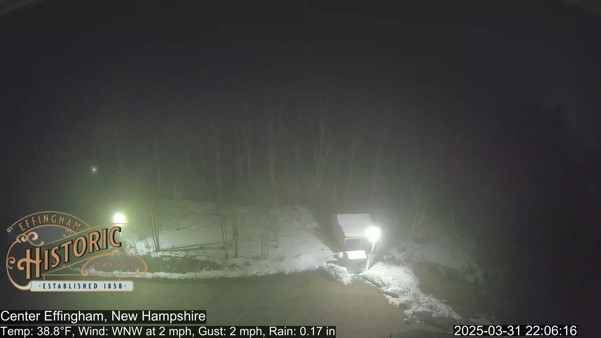 A nighttime view shows a snow-covered area with a small structure and light posts under foggy conditions and light rain.