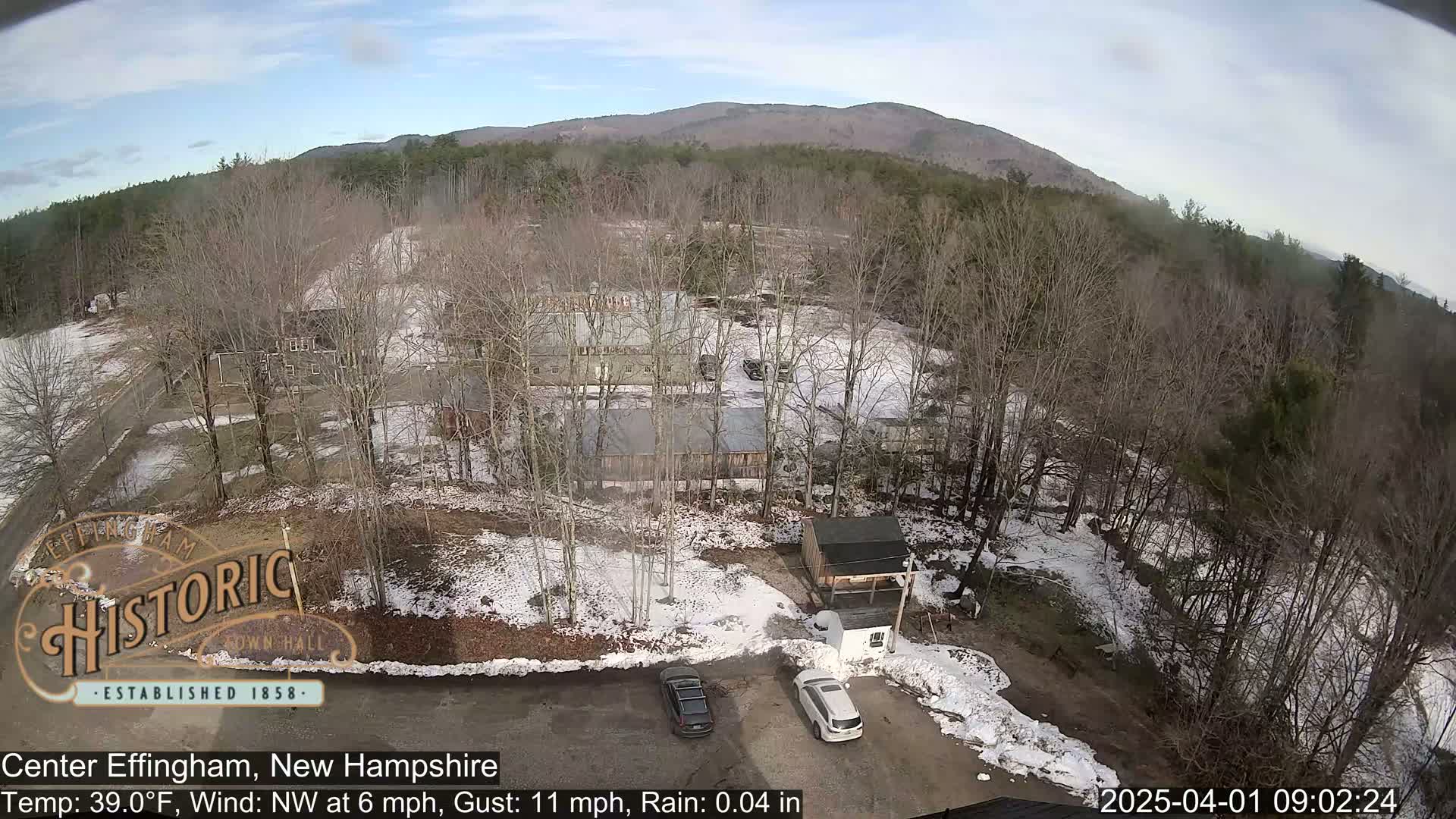 A high-angle view shows a snow-dusted landscape with several buildings, two cars in a parking lot, and bare trees under a partly cloudy sky.