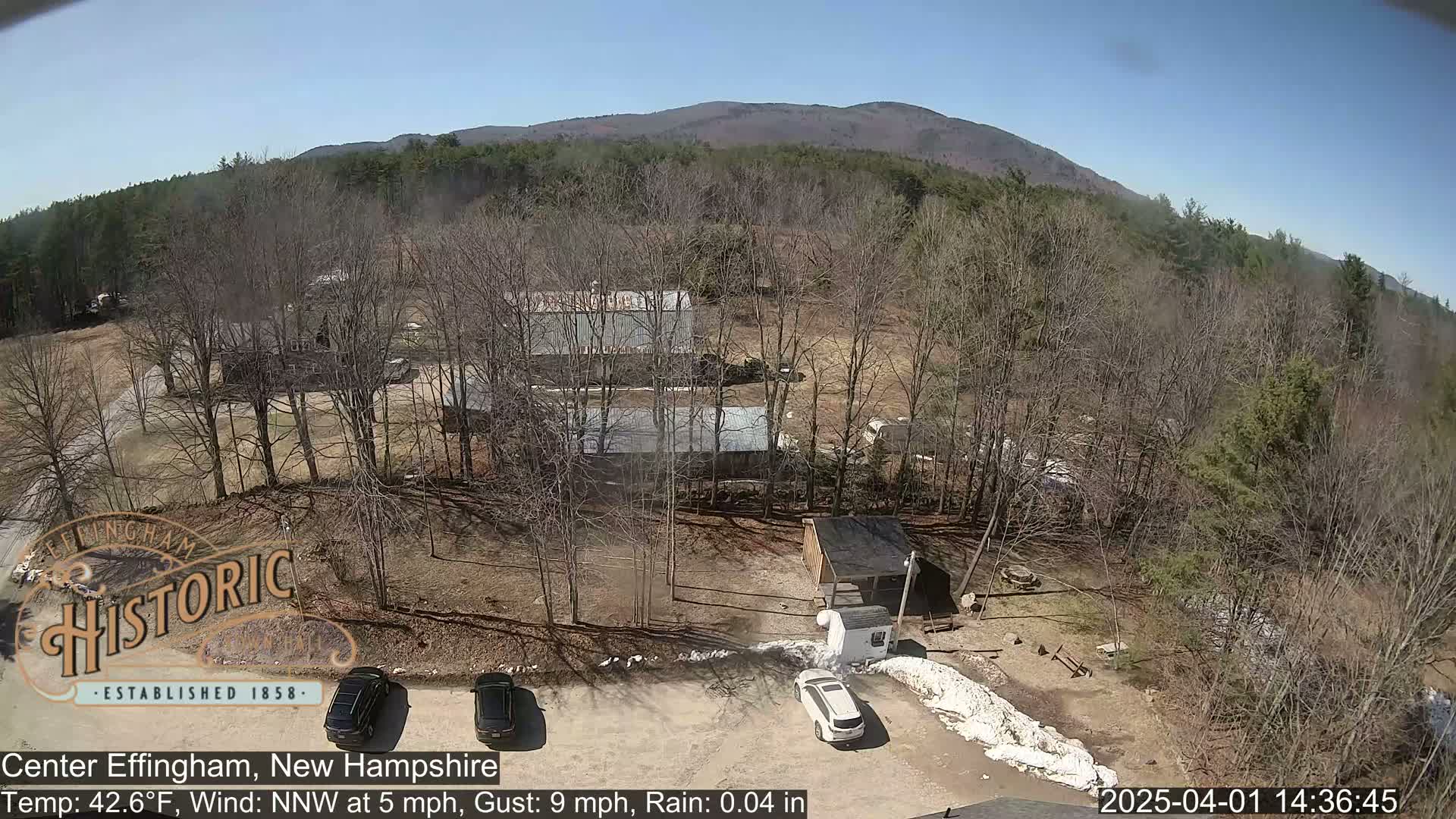 An aerial view shows a parking lot with two cars, several buildings surrounded by bare trees in a lightly wooded area under a sunny sky, with a mountain in the background.