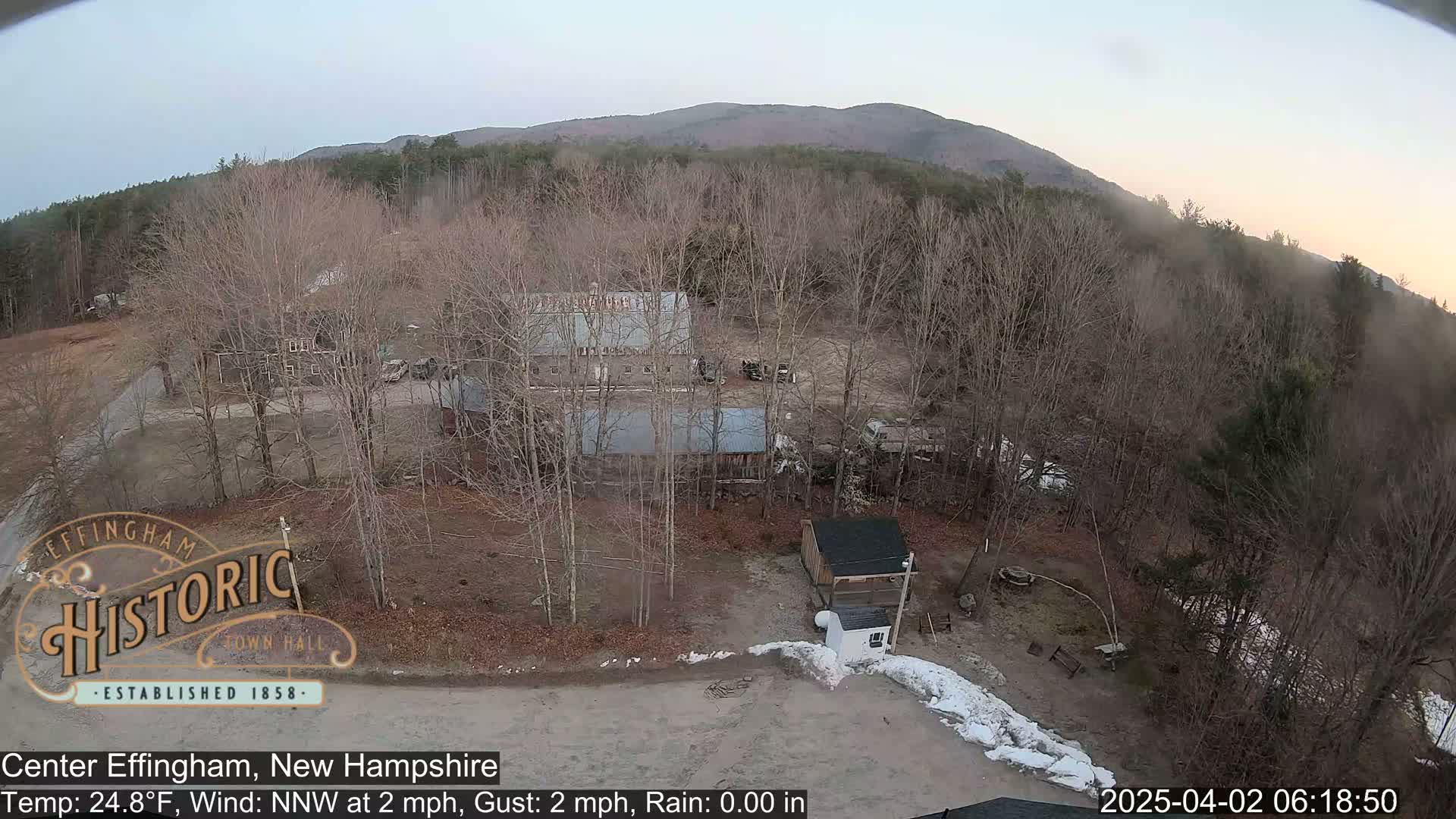 An aerial view shows a rural area with several buildings, bare trees, and a mountain in the background under a clear, dusky sky.