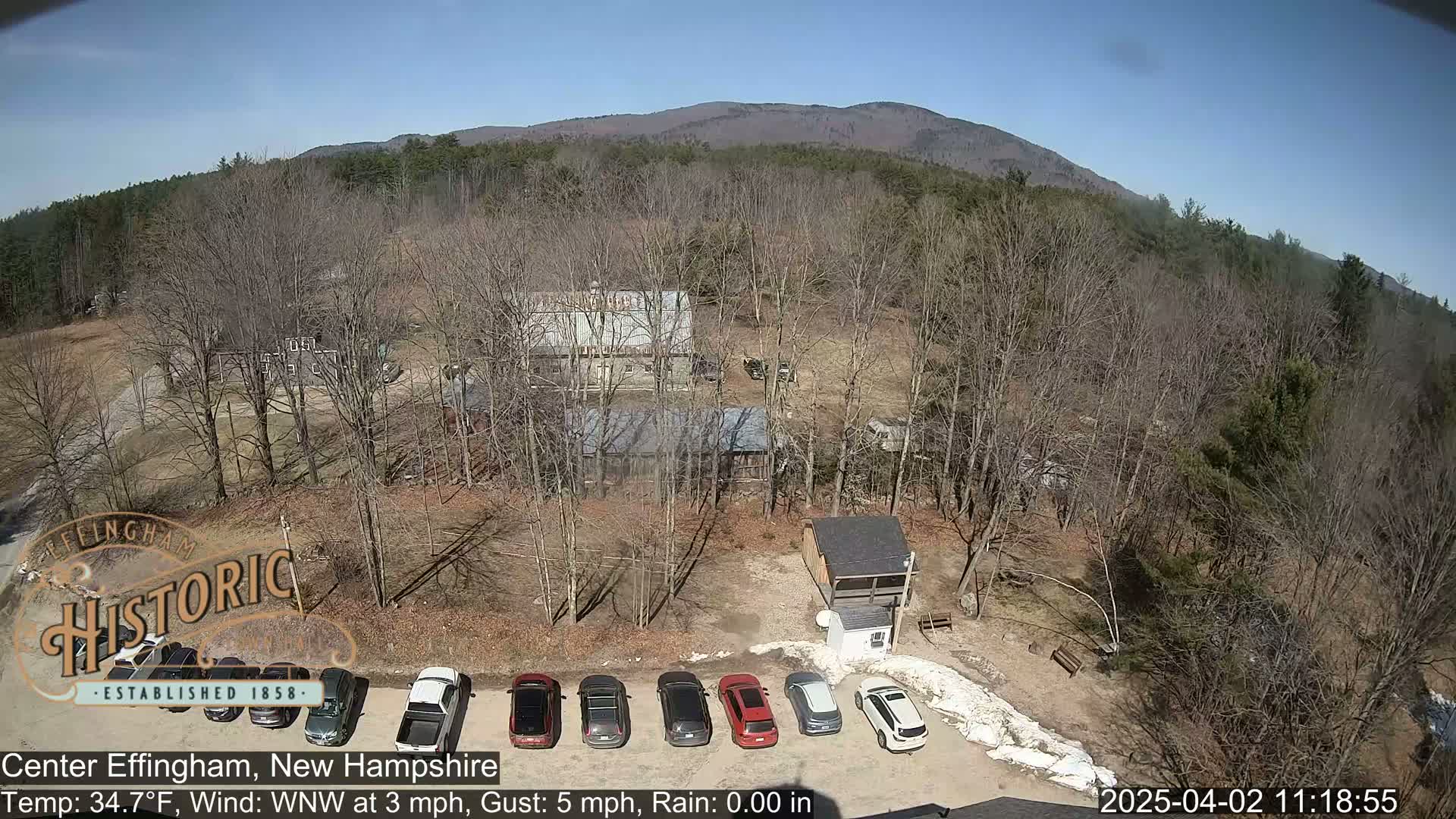 A parking lot with several cars is situated near several buildings in a wooded area with a mountain in the background on a clear, sunny day.